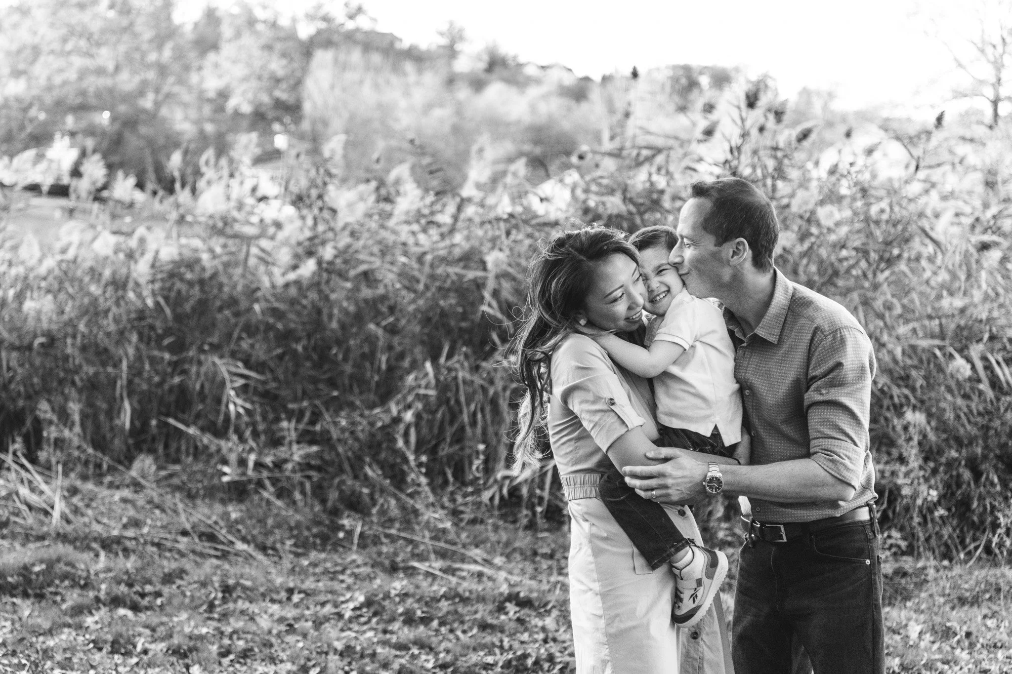  A father kisses his little boy in his mother's arms by Nicole Hawkins Photography. black and white family portrait #NicoleHawkinsPhotography #NicoleHawkinsFamilies #fallfamilyphotos #NorthernNewJerseyPhotography #outdoorfamilyportraits 