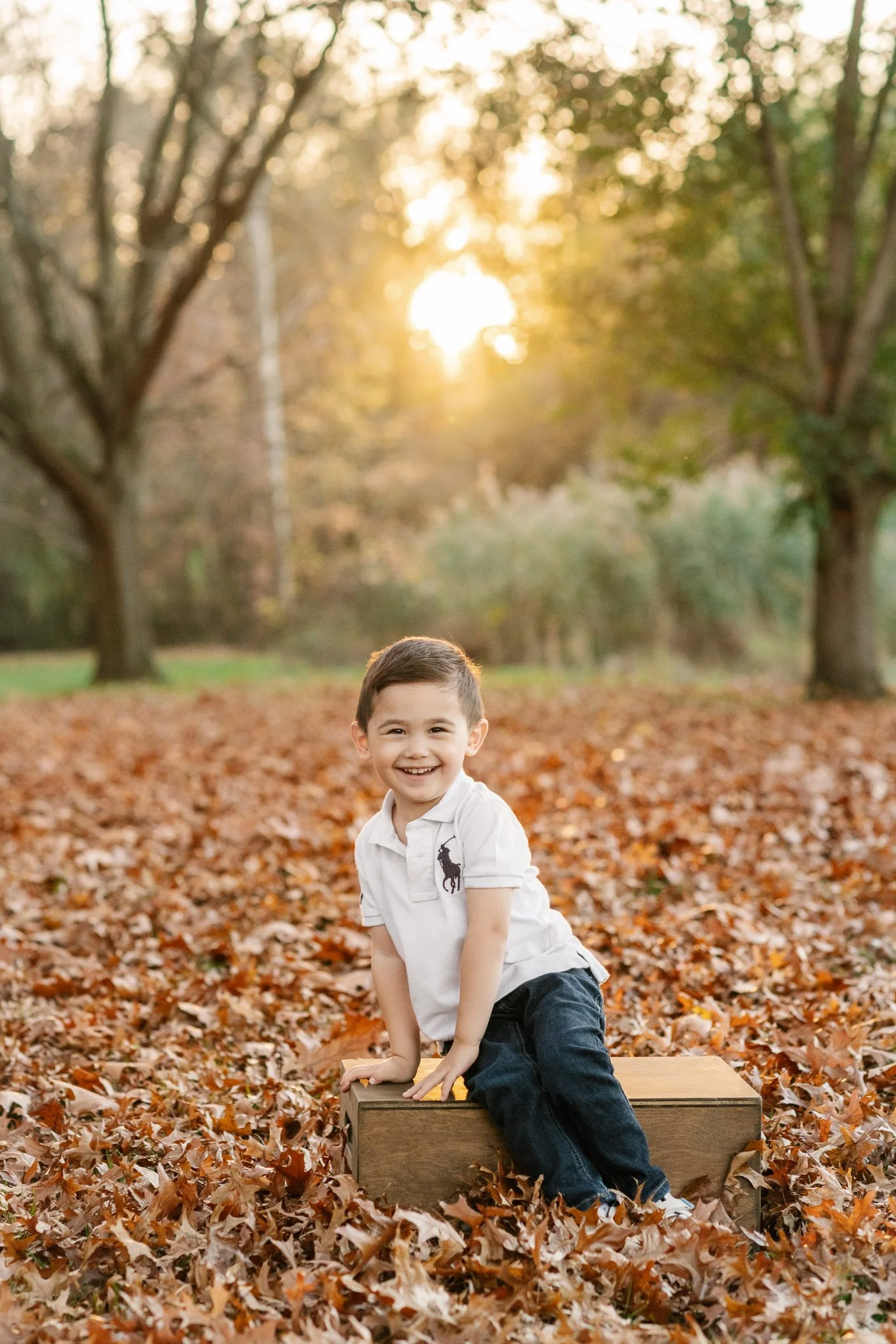  Sitting on a wooden box in a pile of orange leaves a little boy smiles captured by Nicole Hawkins Photography. autumn pics #NicoleHawkinsPhotography #NicoleHawkinsFamilies #fallfamilyphotos #NorthernNewJerseyPhotography #outdoorfamilyportraits 