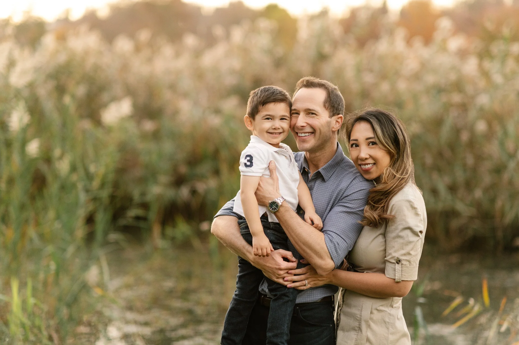  Next to yellow grass, a family of three poses for a family photo by Nicole Hawkins Photography. biracial family #NicoleHawkinsPhotography #NicoleHawkinsFamilies #fallfamilyphoto #NorthernNewJerseyPhotography #outdoorfamilyportraits 