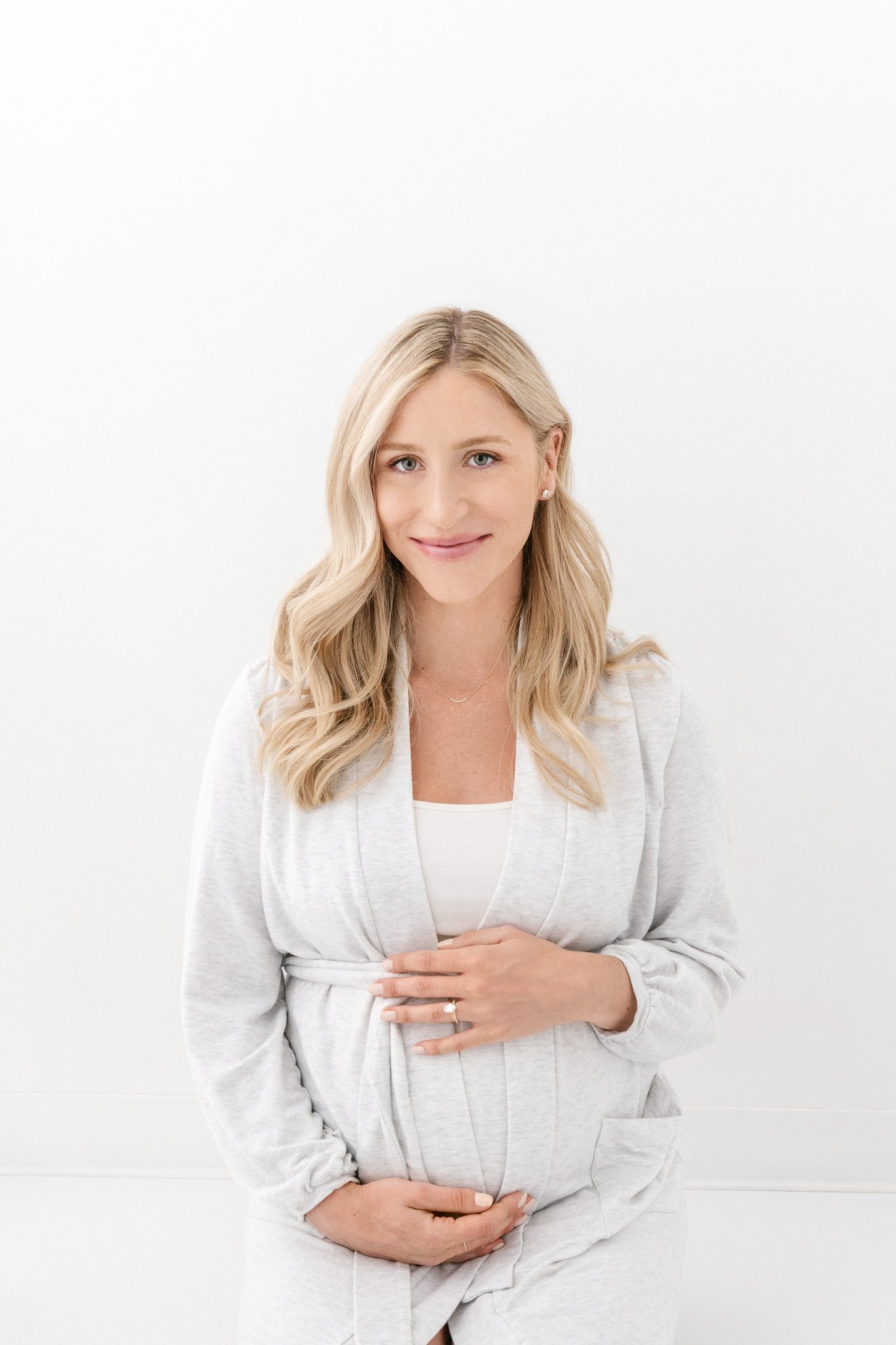  A blonde woman holds her pregnant baby bump as she smiles by Nicole Hawkins Photography in NJ. white maternity dress #NicoleHawkinsPhotography #NicoleHawkinsMaternity #Studiomaternityportraits #NewJerseymaternity #NewYorkMaternity 