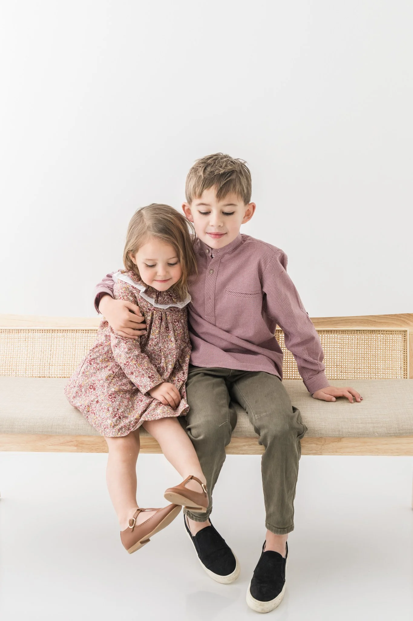  Two children swing their legs on a rattan bench in a New Jersey studio by Nicole Hawkins Photography. rattan bench photos #NicoleHawkinsPhotography #NicoleHawkinsFamily #Newborns #MaplewoodNJ #studiofamilyportraits #modernfamilyportraits 
