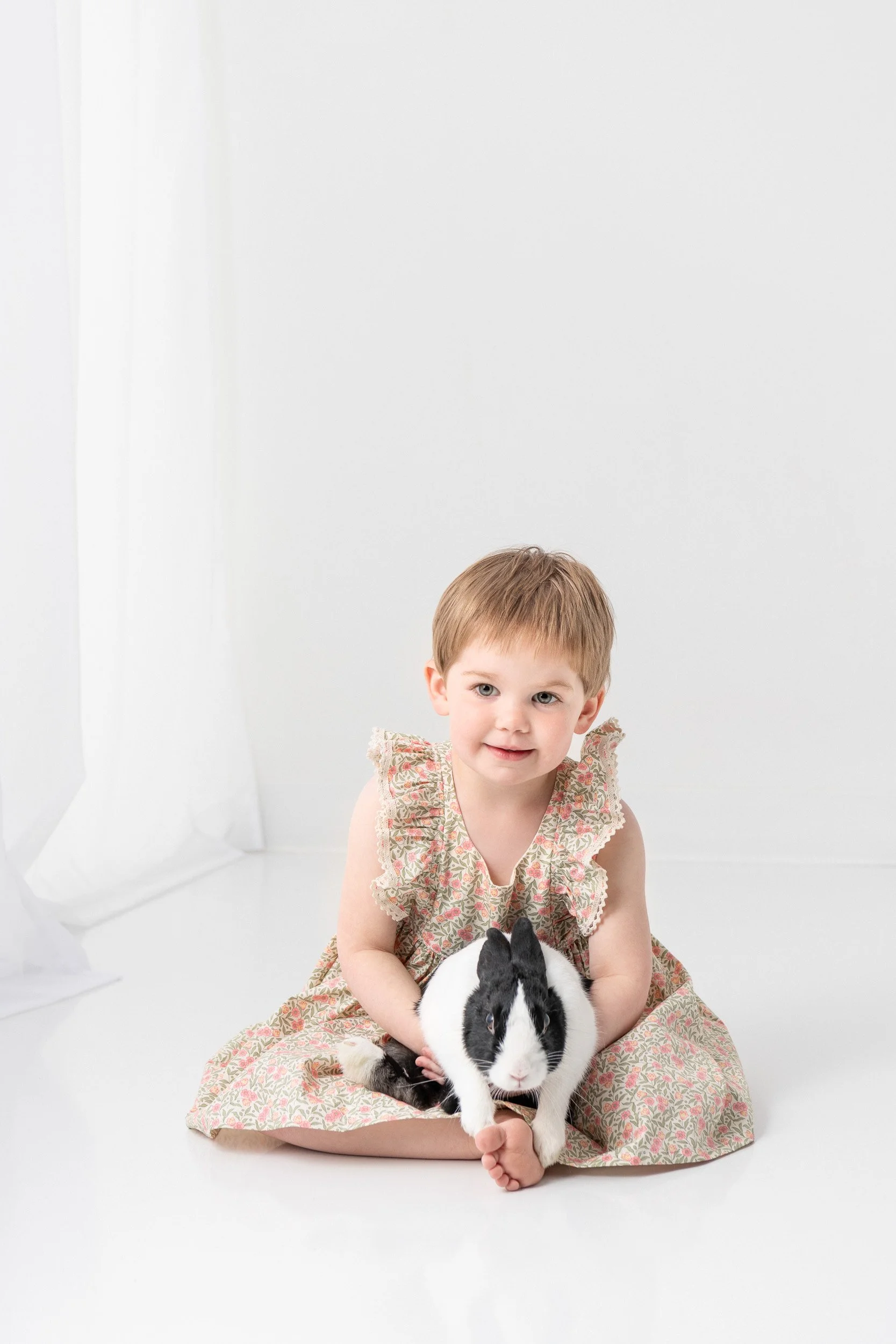  A toddler girl holds a black and white bunny in a floral dress by Nicole Hawkins Photography. toddler girl with short hair #NicoleHawkinsPhotography #NicoleHawkinsChildren #3rdbirthday #studioportrait #agirlandherbunny #littlegirl  