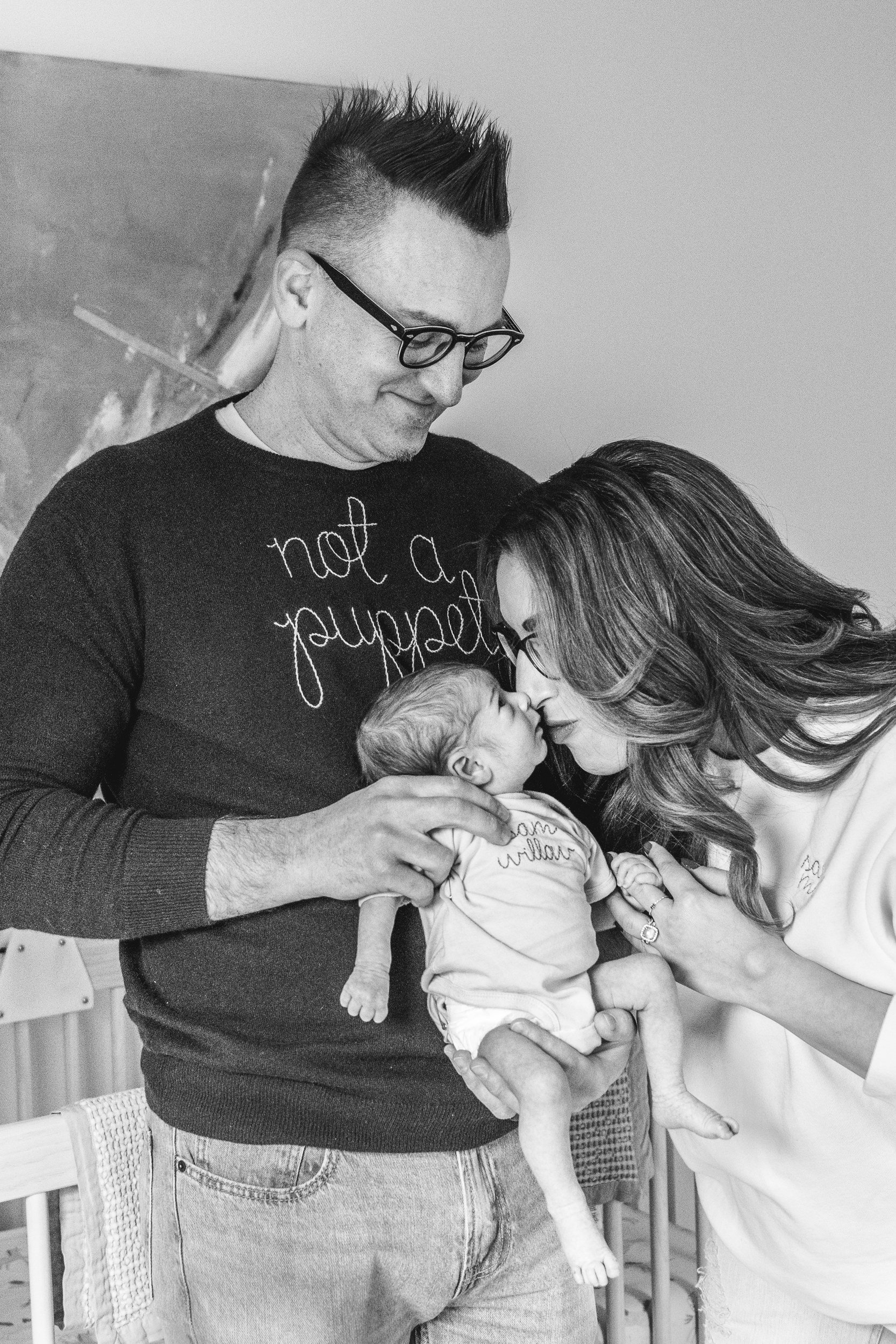  While a dad holds his baby girl a mother leans in to kiss the baby captured by Nicole Hawkins Photography. family of three #NicoleHawkinsPhotography #NicoleHawkinsFamily #Newborns #InHomeNewborns #NicoleHawkinsNewborns #NJnewborns #babygirl 