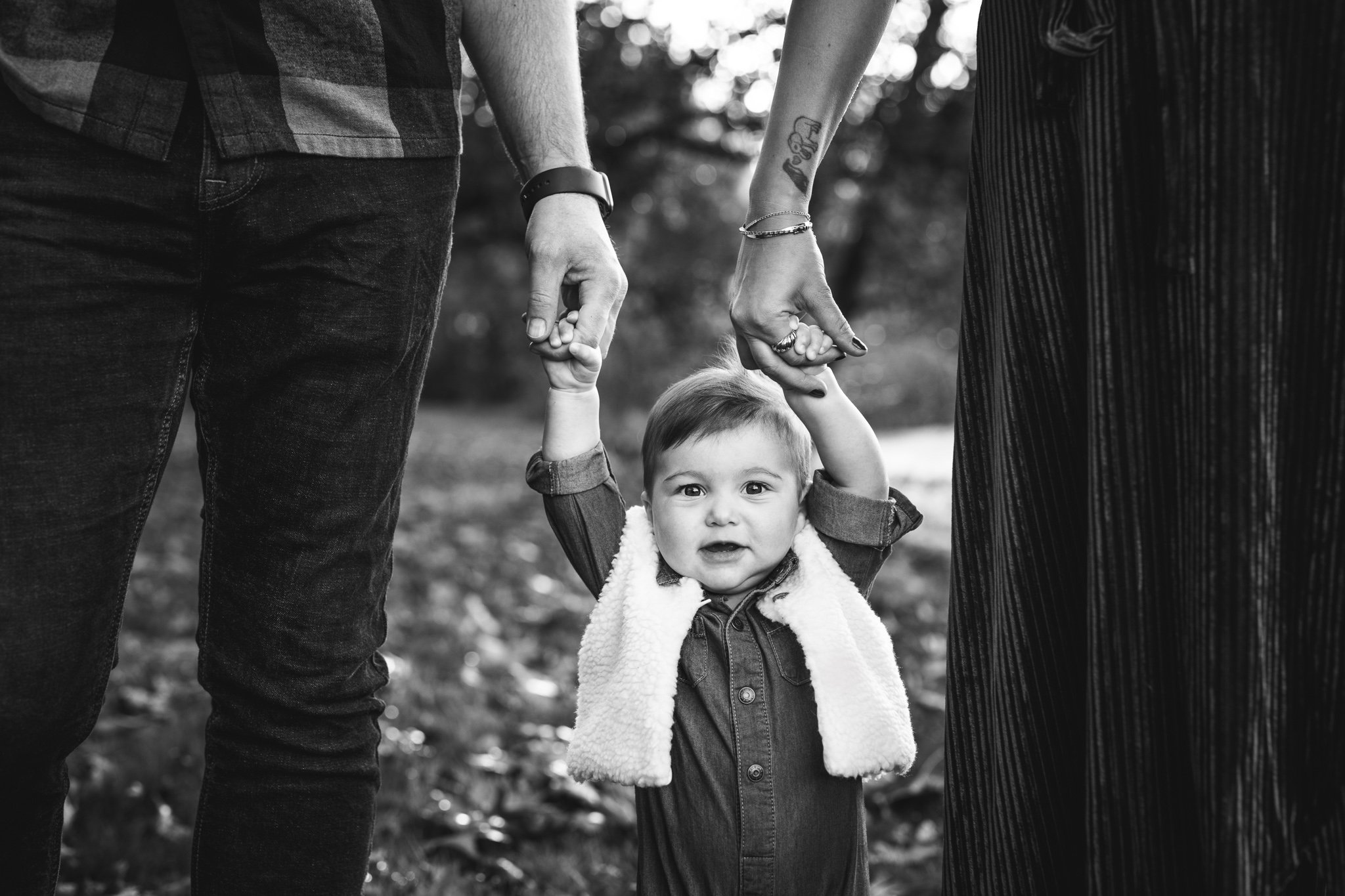  Black and white edit of a baby walking while holding mom and dad's hands by Nicole Hawkins Photography. NJ family photographer #NicoleHawkinsPhotography #NicoleHawkinsFamily #fallfamilyphotos #fallphotos #familyphotographerNJ #NJfallfamilypics 
