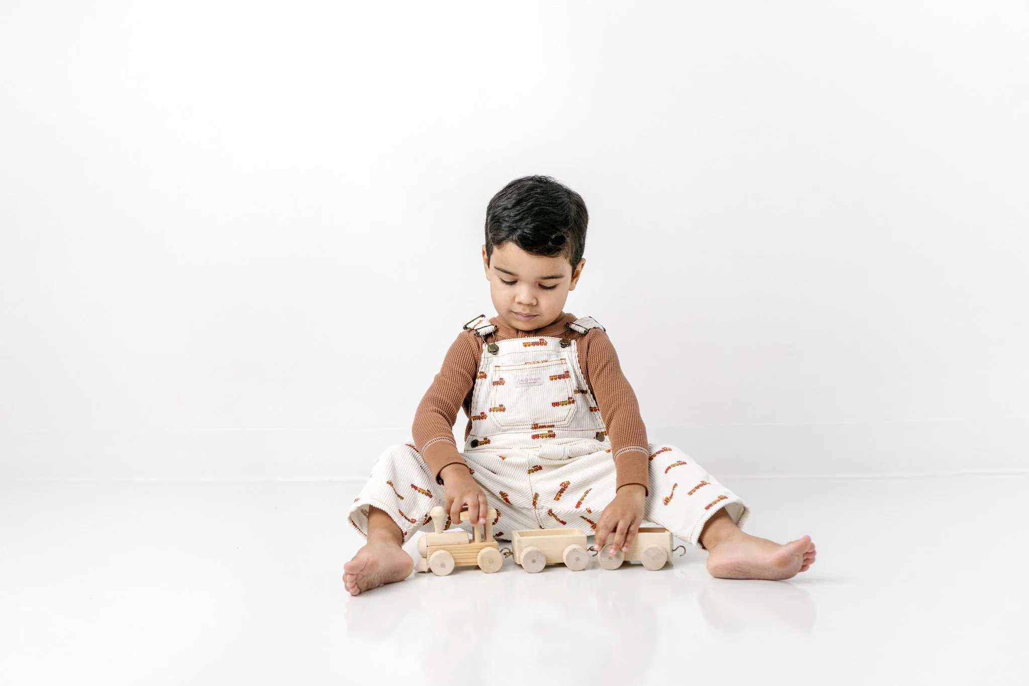  A toddler boy in overalls plays with a wooden train during a studio session with Nicole Hawkins Photography. modern studio #NicoleHawkinsPhotograaphy #NicoleHawkinsChildren #StudioBabies&amp;Children #NJphotographers #modernportraits #mom&amp;meport