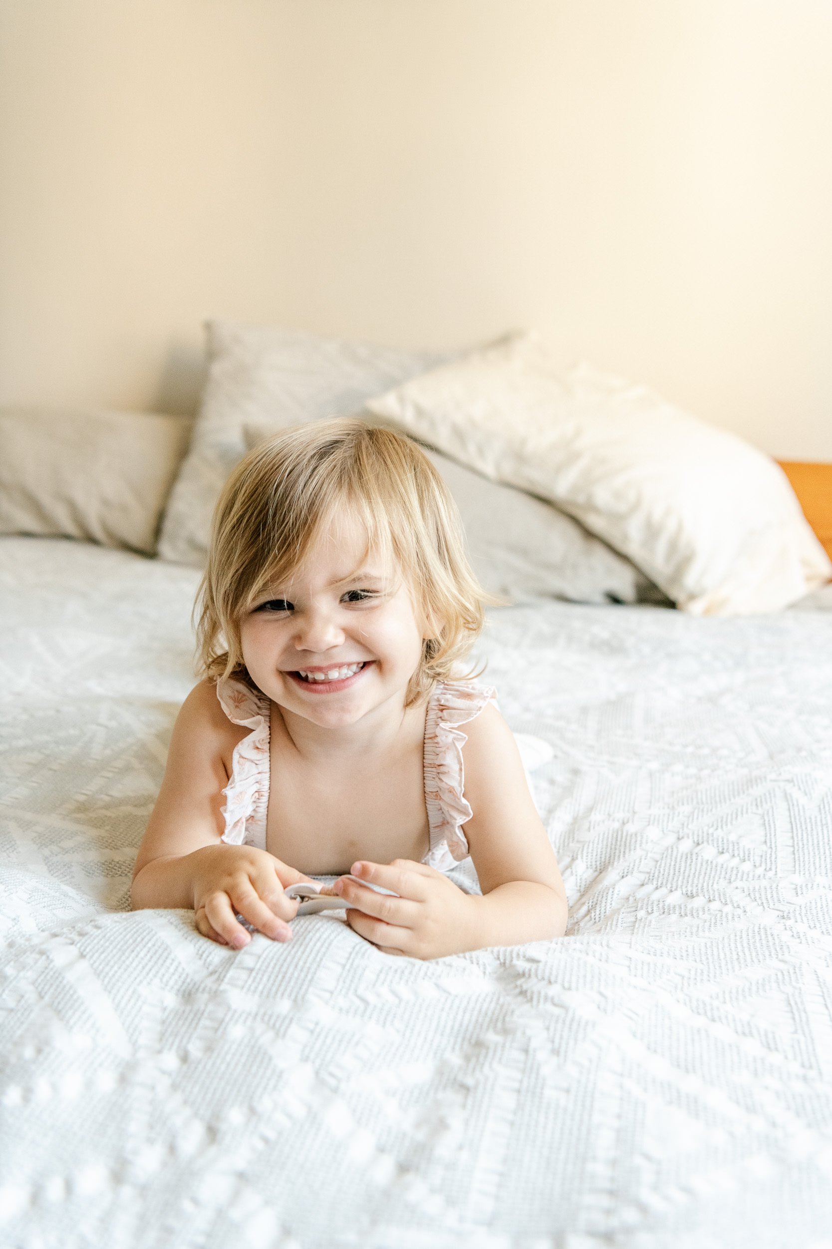  Little girl on a white bed laying on her belly for an at-home portrait by Nicole Hawkins Photography. toddler bed photos #NicoleHawkinsPhotograaphy #NicoleHawkinsChildren #NewYorkPortraits #KidsinNewYork #ChildrensPortraits #lifestyleportraits 