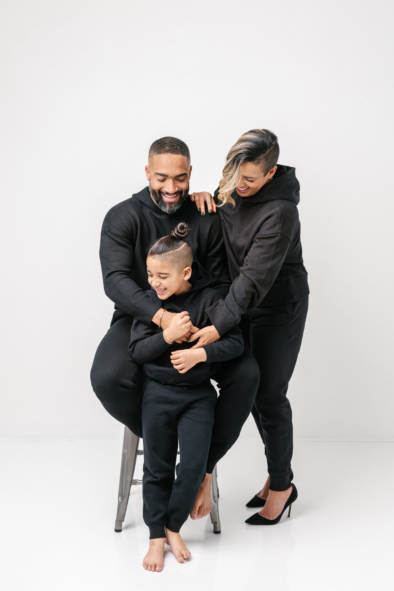  A little boy sits on a stool while his mother and father hug him by Nicole Hawkins Photography. family with boy #NicoleHawkinsPhotograaphy #NicoleHawkinsFamilies #Modernfamilypictures #studioportraits #matchingfamilyoutfits #NJphotographer 