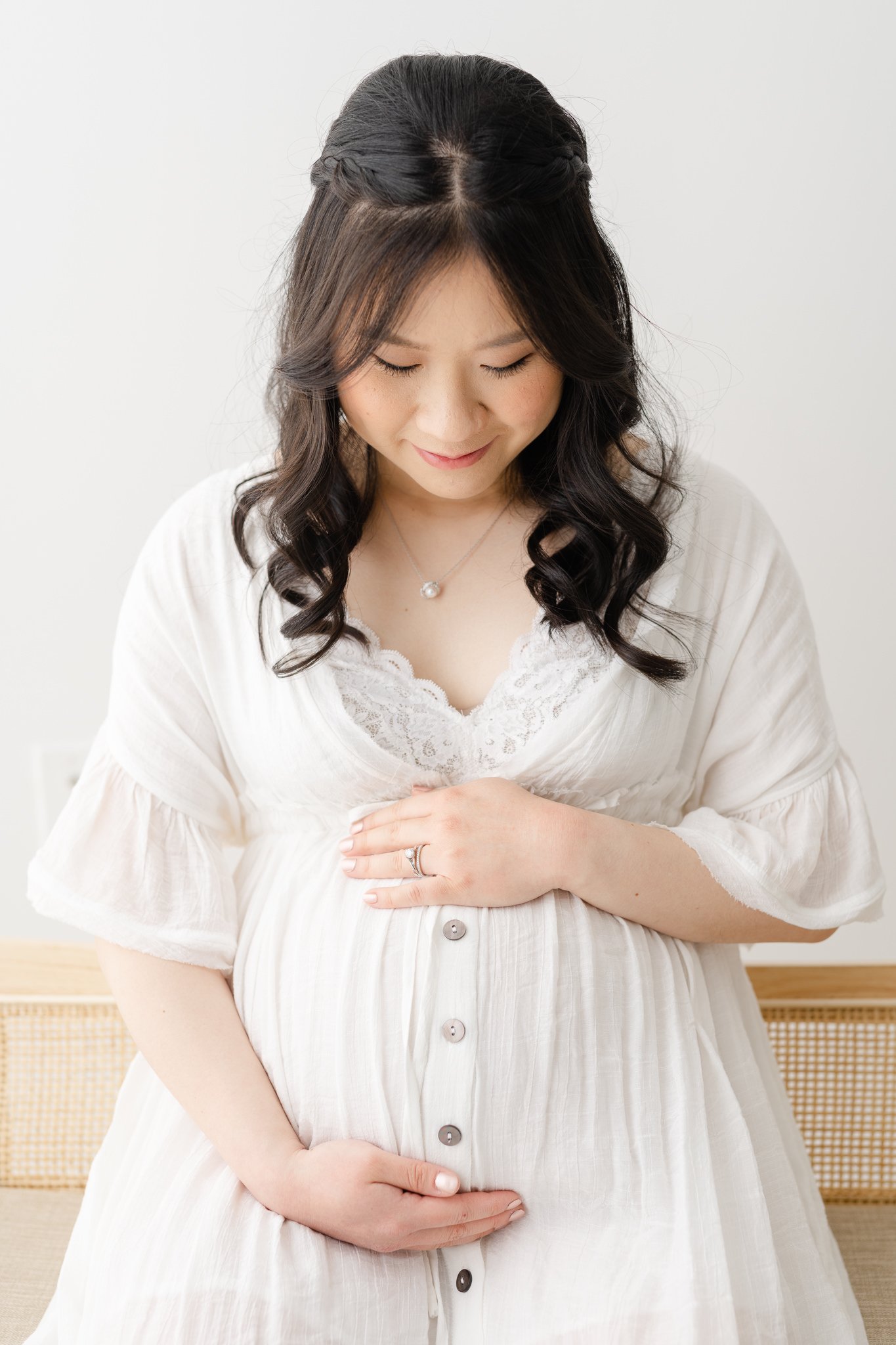  A close-up portrait of an Asian mother holding her baby bump in studio maternity portraits with Nicole Hawkins Photography. white maternity dress outfit inspiration #NicoleHawkinsPhotography #NicoleHawkinsMaternity #MaternityPhotography #Maternityst