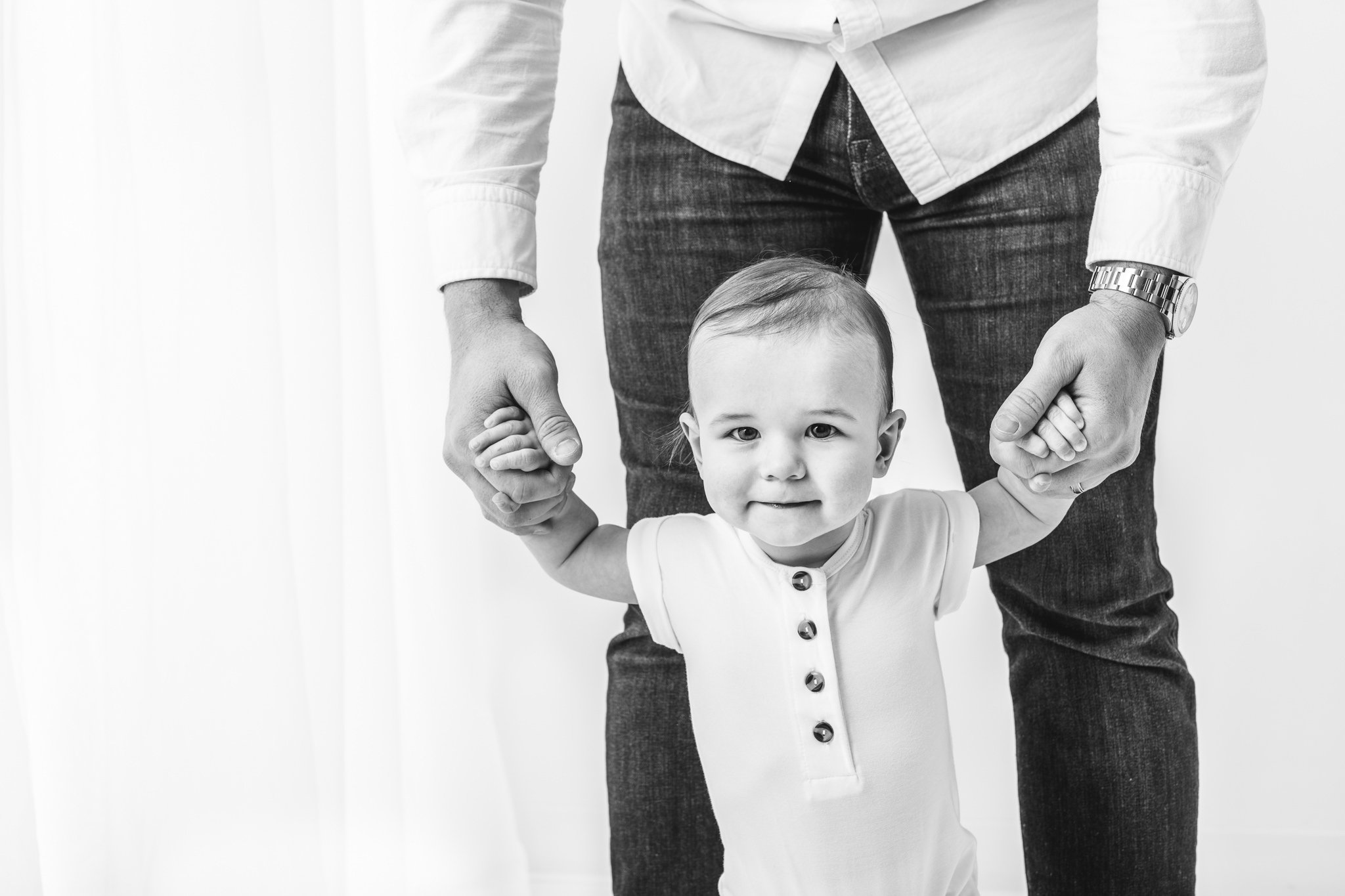  A black and white portrait of a one-year-old boy walking while holding his daddy's hands by Nicole Hawkins PHotography. walking baby boy timeless classic baby portraits #NicoleHawkinsPhotography #NicoleHawkinsBabies #FirstBirthdayPhotography #Studio