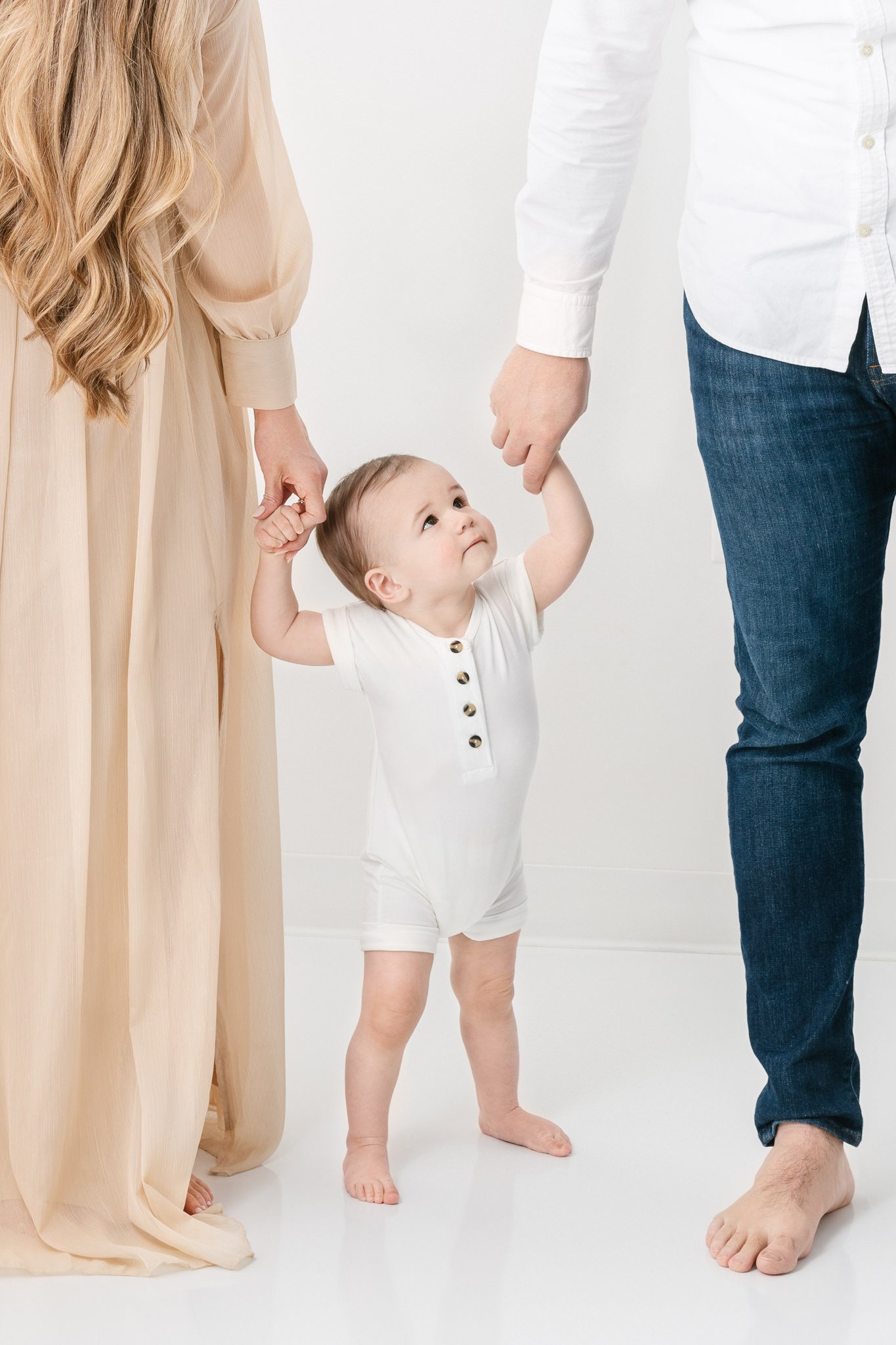  Toddler baby holds his parent's hands and looks up at them captured by Nicole Hawkins Photography in New Jersey. toddler poses with parents romper for boy #NicoleHawkinsPhotography #NicoleHawkinsBabies #FirstBirthdayPhotography #StudioFamilyPortrait
