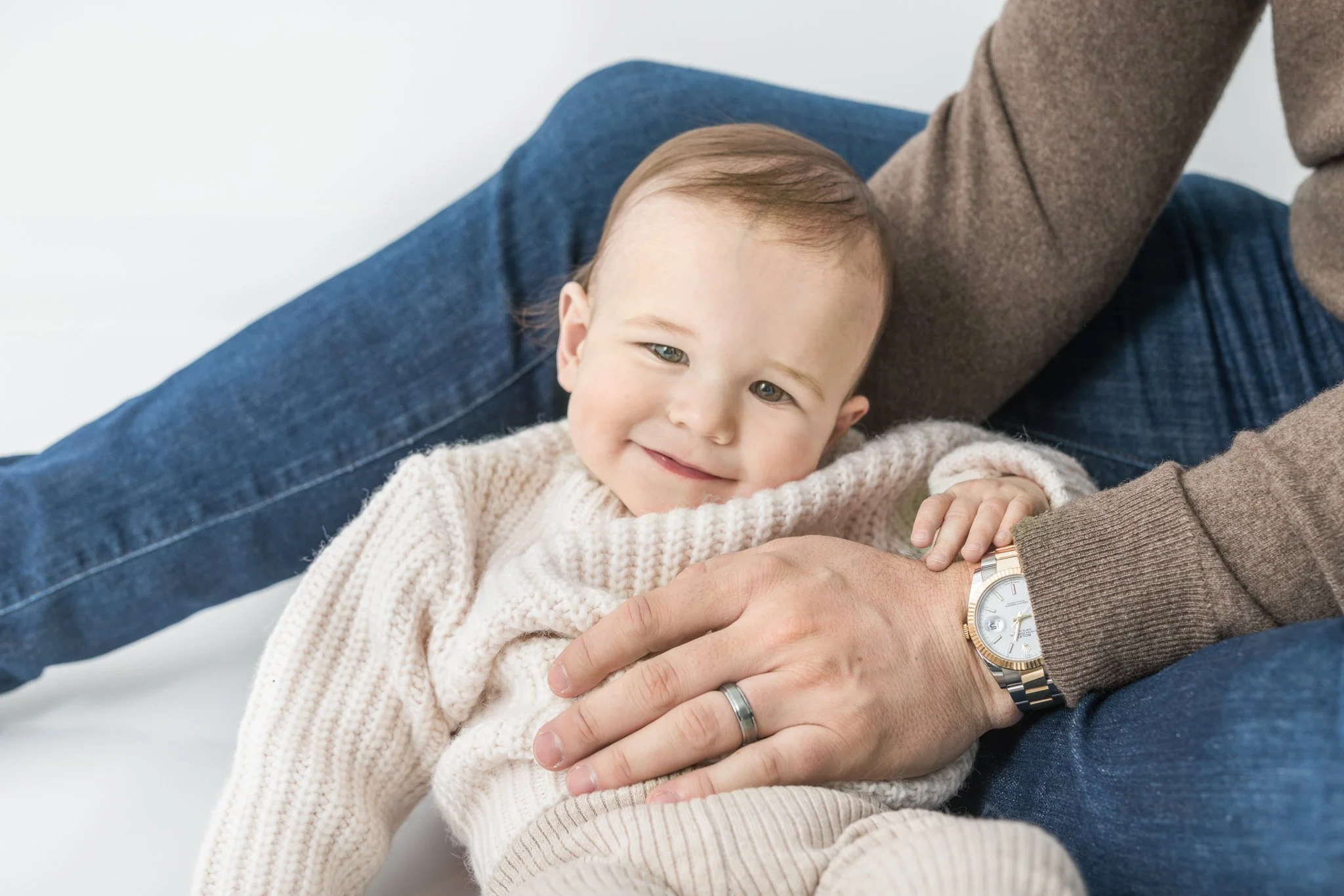  A baby lounges on his father's lap smiling captured by professional studio photographer Nicole Hawkins Photography. studio photographers NJ/NY baby boy outfit inspiration #NicoleHawkinsPhotography #NicoleHawkinsBabies #FirstBirthdayPhotography #Stud