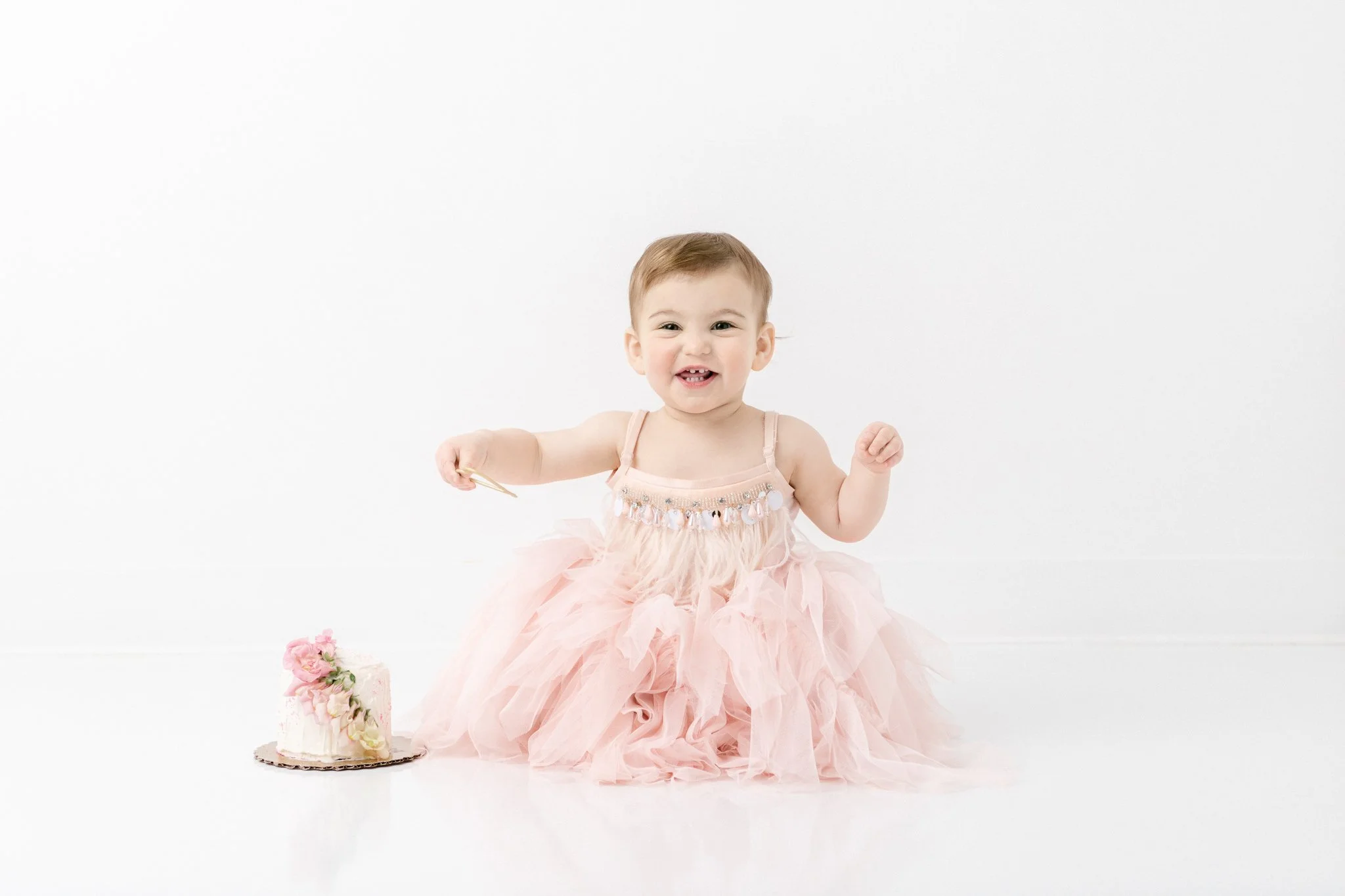  A baby girl in a ruffle dress points to her cake by Nicole Hawkins Photography in a white studio. cute baby girl dress ideas smash cake floral #NicoleHawkinsPhotography #NicoleHawkinsBabies&amp;Children #FirstBirthdayPhotography #StudioPhotography #