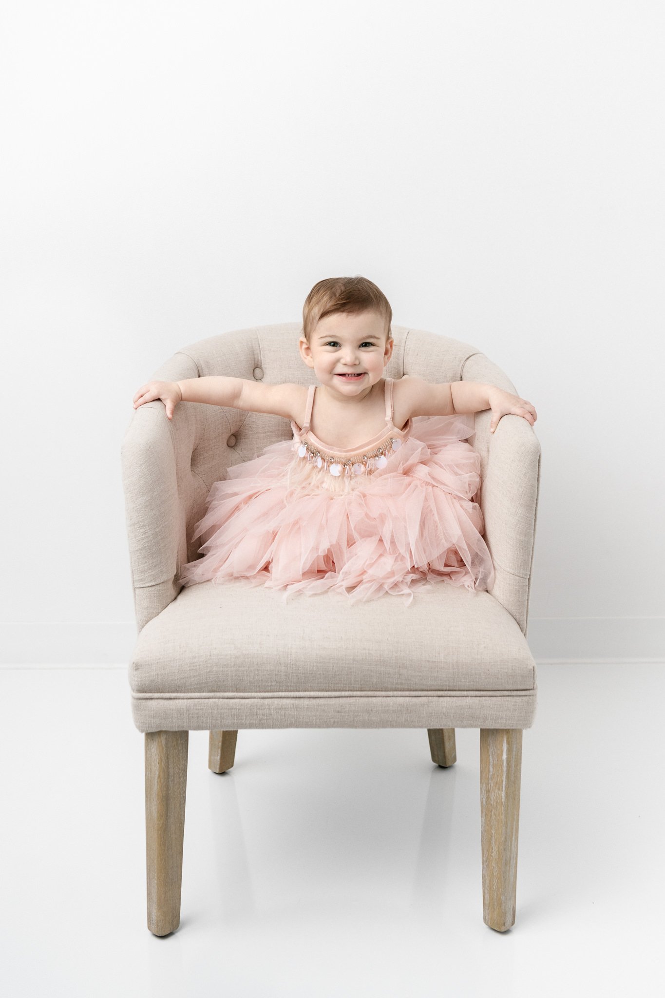  A spunky little girl sits in a chair and smiles during her first birthday session with Nicole Hawkins Photography. spunky baby girl portraits fun baby girl dress #NicoleHawkinsPhotography #NicoleHawkinsBabies&amp;Children #FirstBirthdayPhotography #