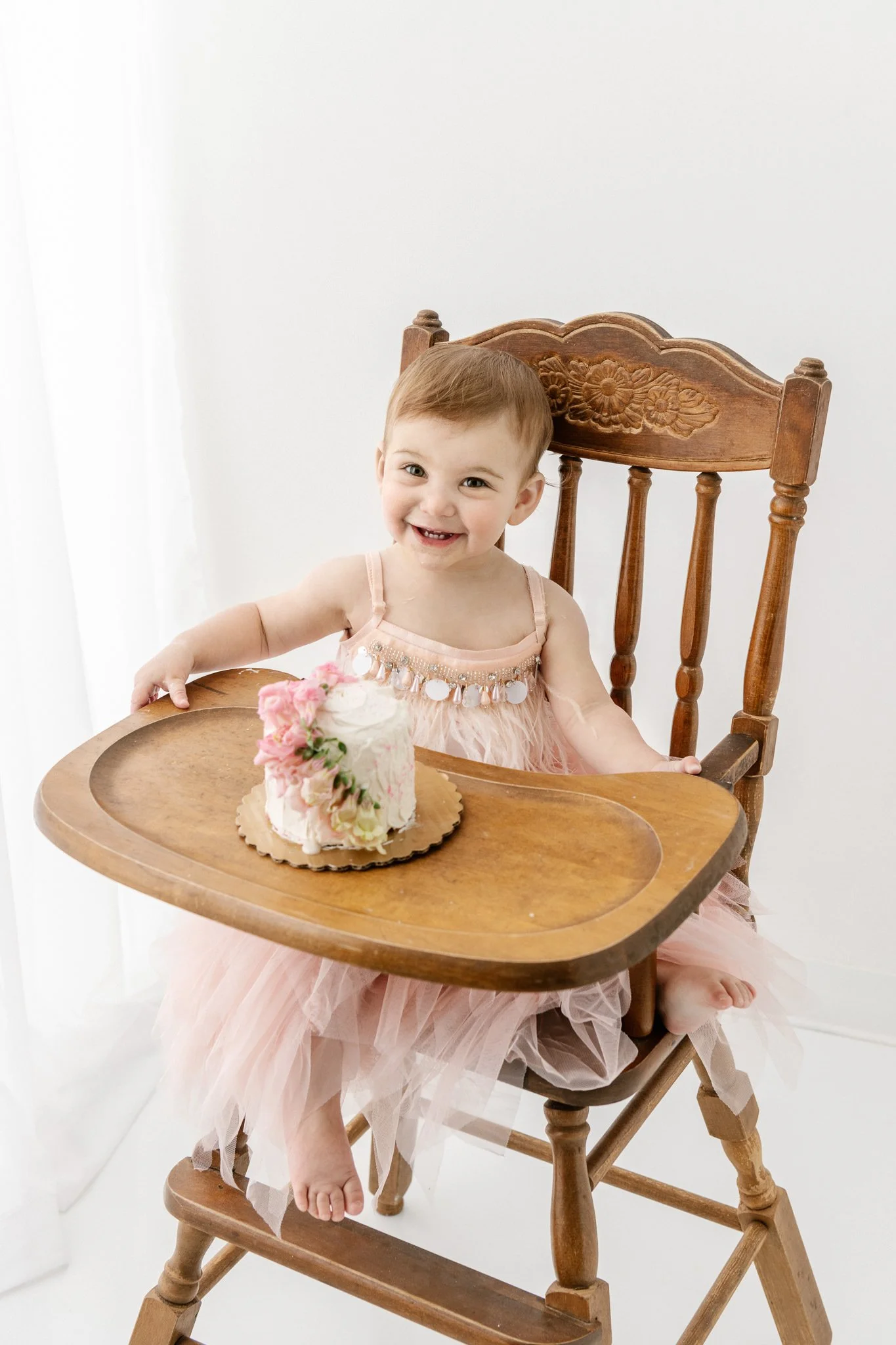  Baby girl sits on a high chair with her floral pink smash cake and smiles by Nicole Hawkins PHotgrpahy in NJ. NJ child photographer NJ studio child photography #NicoleHawkinsPhotography #NicoleHawkinsBabies&amp;Children #FirstBirthdayPhotography #St