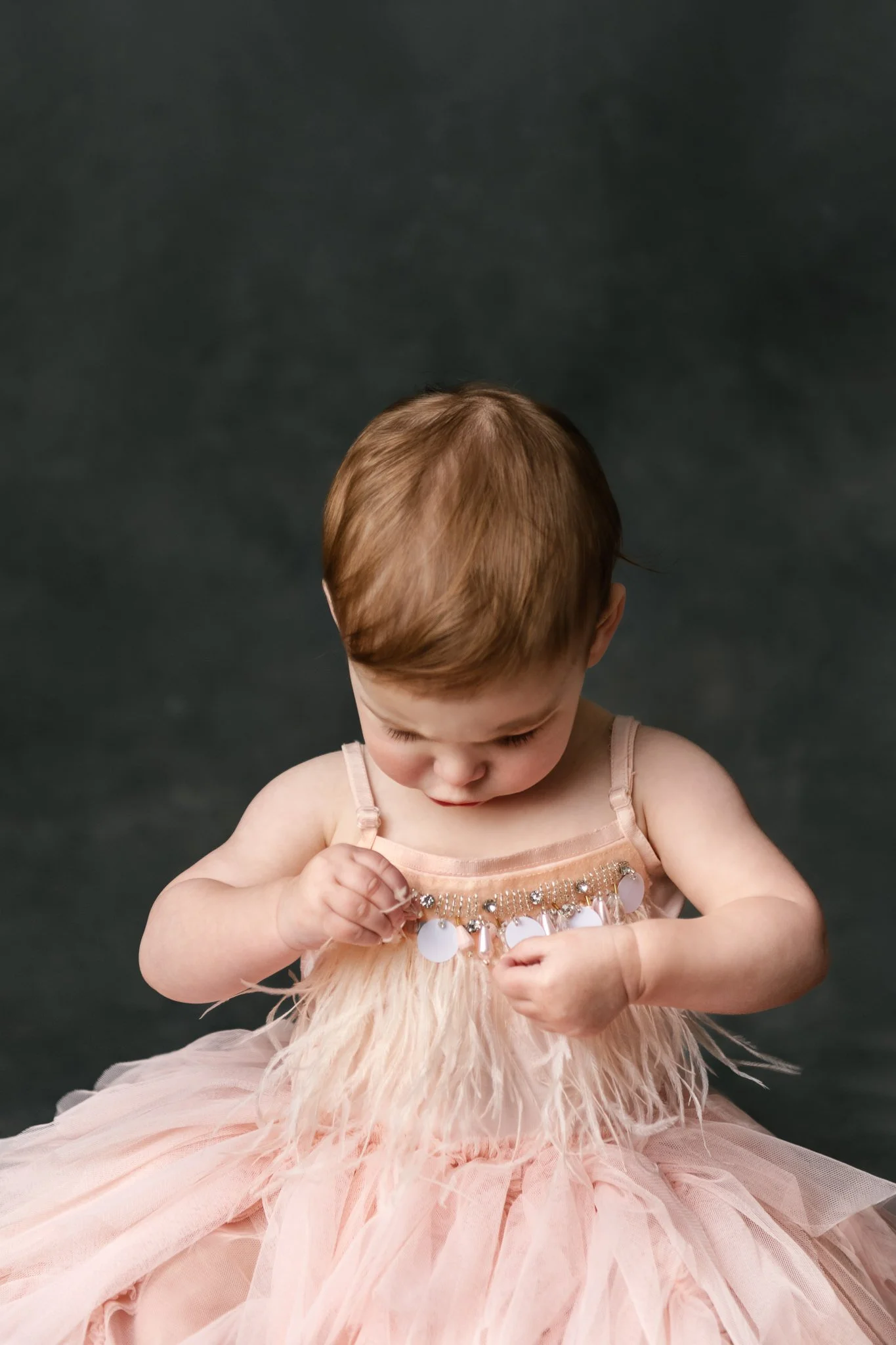  A one-year-old little girl is captured inspecting her birthday tutu dress during studio portraits by Nicole Hawkins Photography. studio portraits flapper dress baby peach dress #NicoleHawkinsPhotography #NicoleHawkinsBabies&amp;Children #FirstBirthd