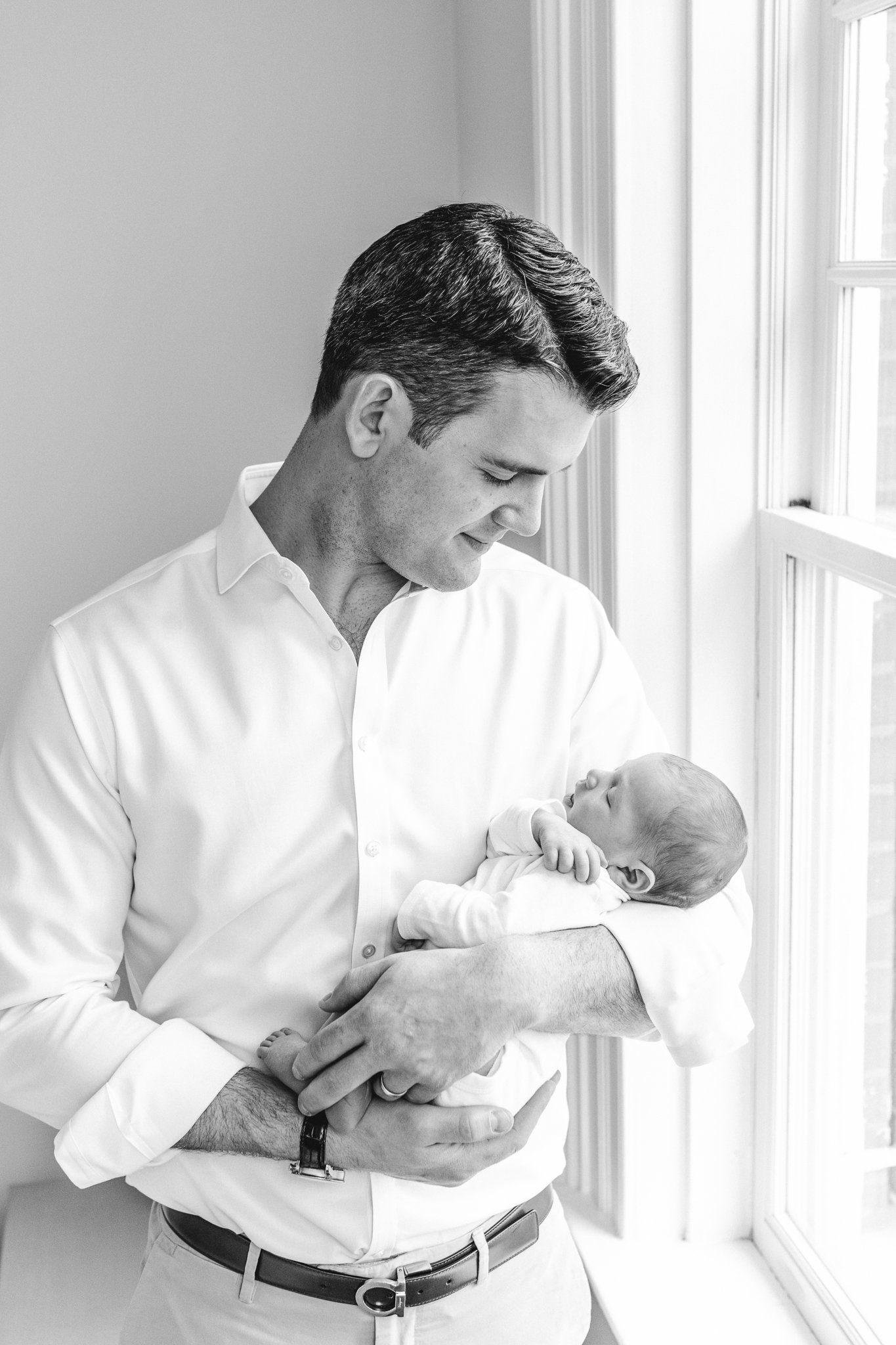  A father holds his baby girl in his arms next to a bright light window during an in-home newborn session in Chatham by Nicole Hawkins Photography. Chatham newborn photographers #NicoleHawkinsPhotography #NicoleHawkinsNewborns #Babygirl #Newbornfamil