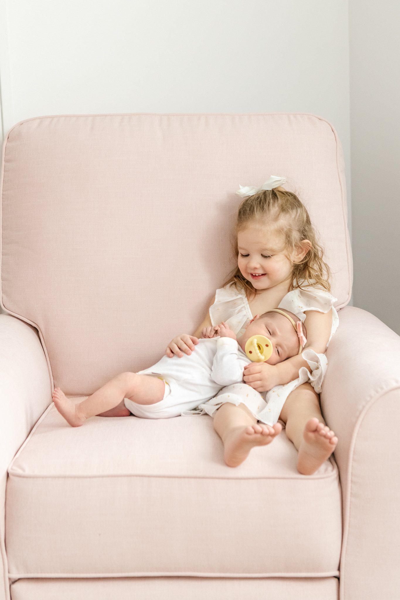  Sitting in a pink rocker a toddler's big sister holds her baby sister in her lap by Nicole Hawkins PHoootgraphy. big sister holding baby sister sisters #NicoleHawkinsPhotography #NicoleHawkinsNewborns #Babygirl #Newbornfamilyportraits #ChathamNewbor
