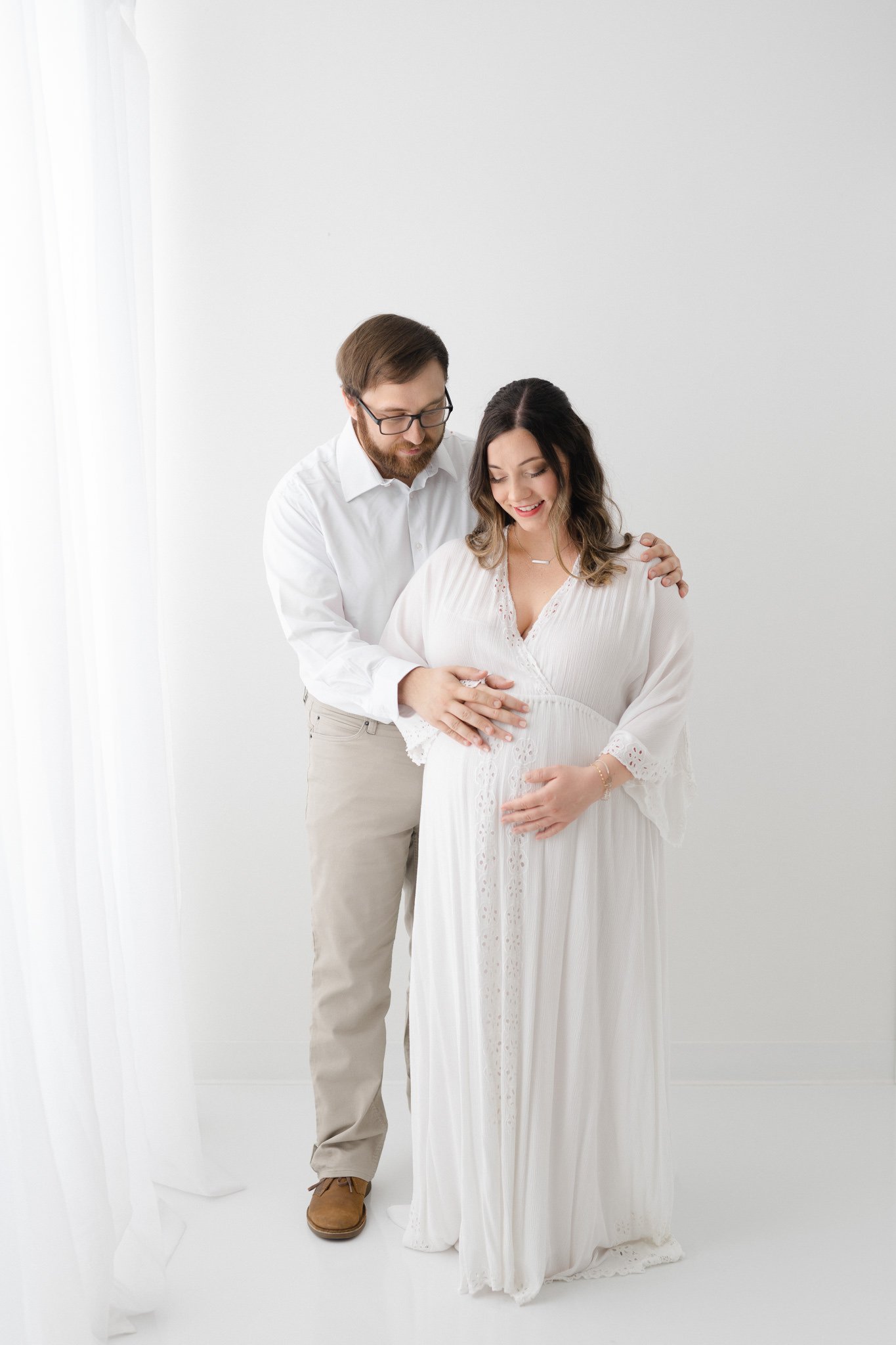  A dad wearing a white shirt and tan pants feels the baby kicking by Nicole Hawkins Photography. feel a baby kick white studio #NicoleHawkinsPhotography #NicoleHawkinsMaternity #Babyontheway #studiomaternityNJ #whitestudiomaternityportraits #NYmatern