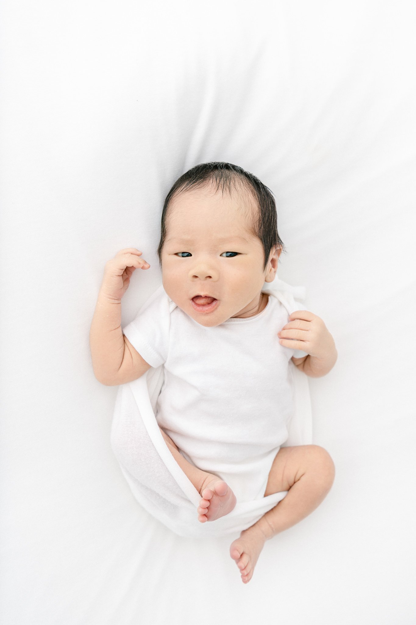  A newborn photographer Nicole Hawkins photography captures a baby boy in a white onesie laying on a bed. baby boy on a bed studio newborns east coast #NicoleHawkinsPhotography #NicoleHawkinsNewborns #Babyboy #studionewbornsNJ #studionewbonportraits 