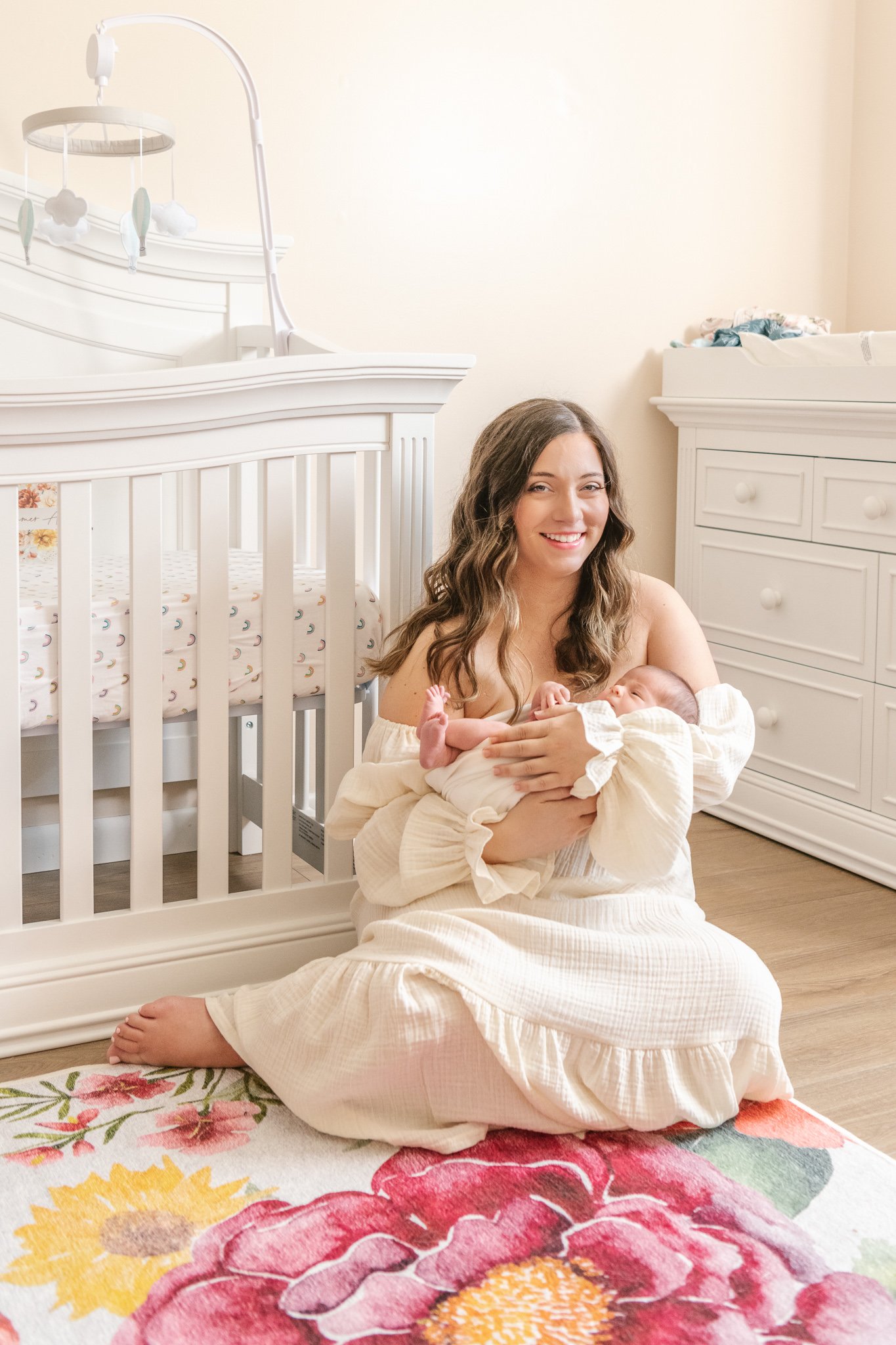  A new mother smiles as she holds her baby girl in her arms sitting on the floor of her nursery by Nicole Hawkins Photography. mother sitting in nursery #NicoleHawkinsPhotography #NicoleHawkinsNewborns #Babygirl #inhomenewbornsNJ #homenewbonportraits