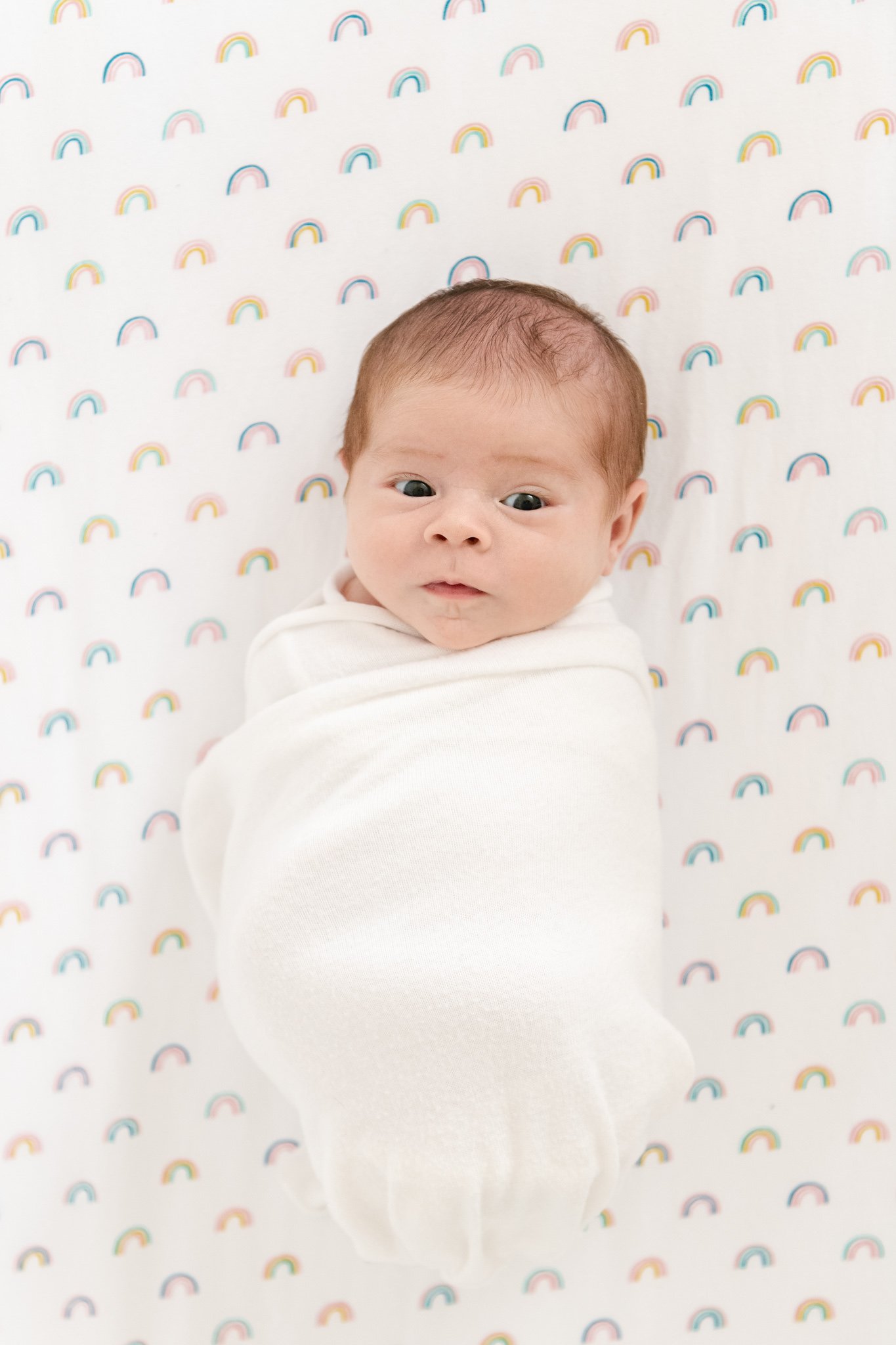  A baby girl lays swaddled up in her crib at her home during a newborn session with Nicole Hawkins Photography. baby on rainbow sheets swaddled baby girl #NicoleHawkinsPhotography #NicoleHawkinsNewborns #Babygirl #inhomenewbornsNJ #homenewbonportrait