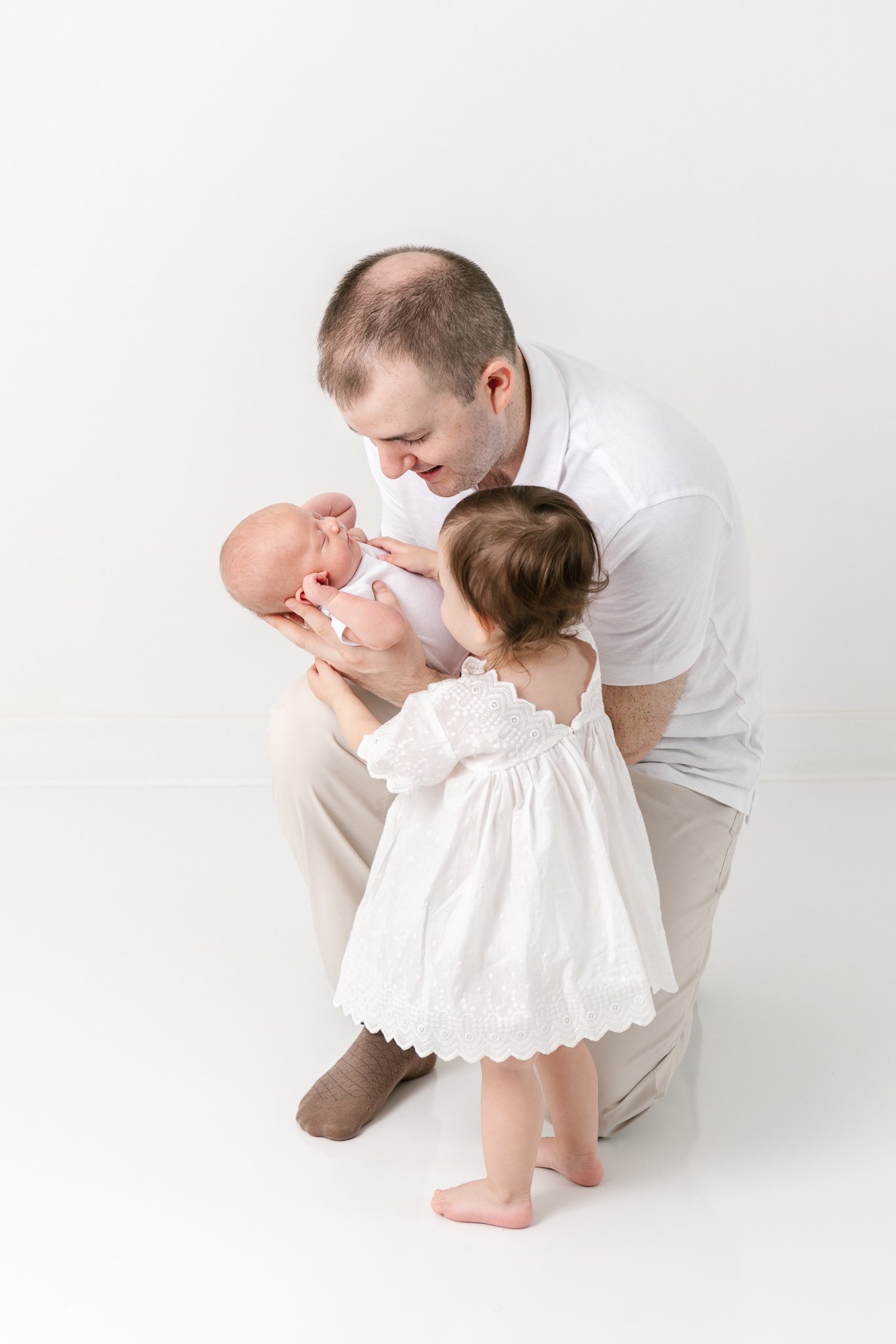  A father holds a newborn daughter showing his toddler little girl her sister by Nicole Hawkins Photography. father and daughter portraits white dress toddler #NicoleHawkinsPhotography #NicoleHawkinsNewborns #StudioNewborn #StudioFamily #EastCoastNew