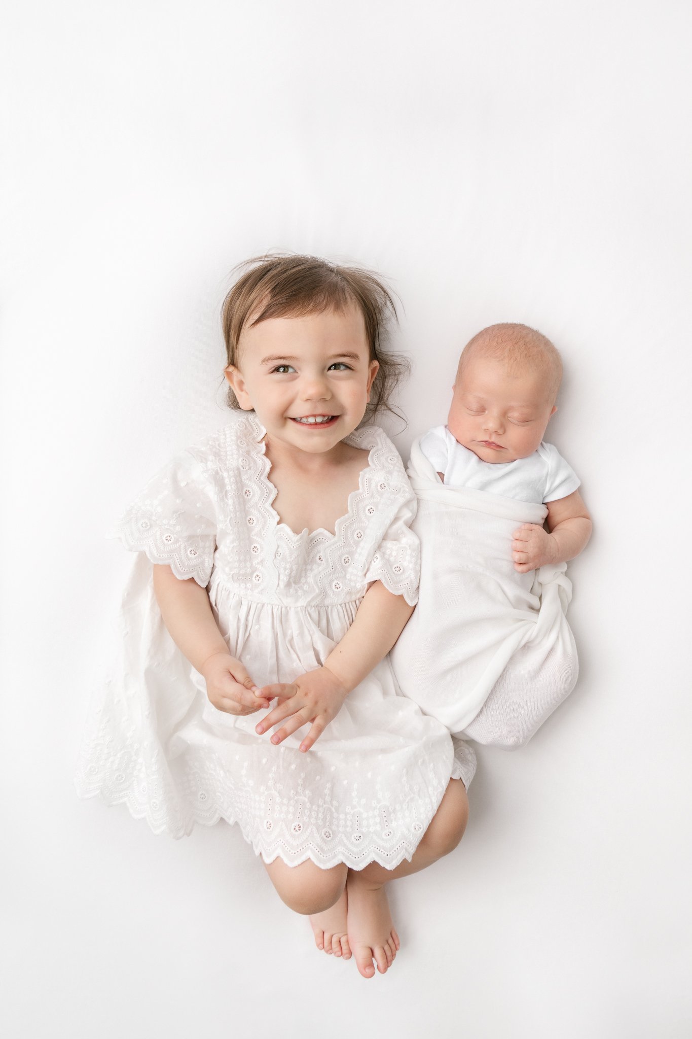 A full body above portrait of two sisters laying on a white bed by Nicole Hawkins Photography. sisters full body portrait timeless sister portrait #NicoleHawkinsPhotography #NicoleHawkinsNewborns #StudioNewborn #StudioFamily #EastCoastNewbornPhotogr