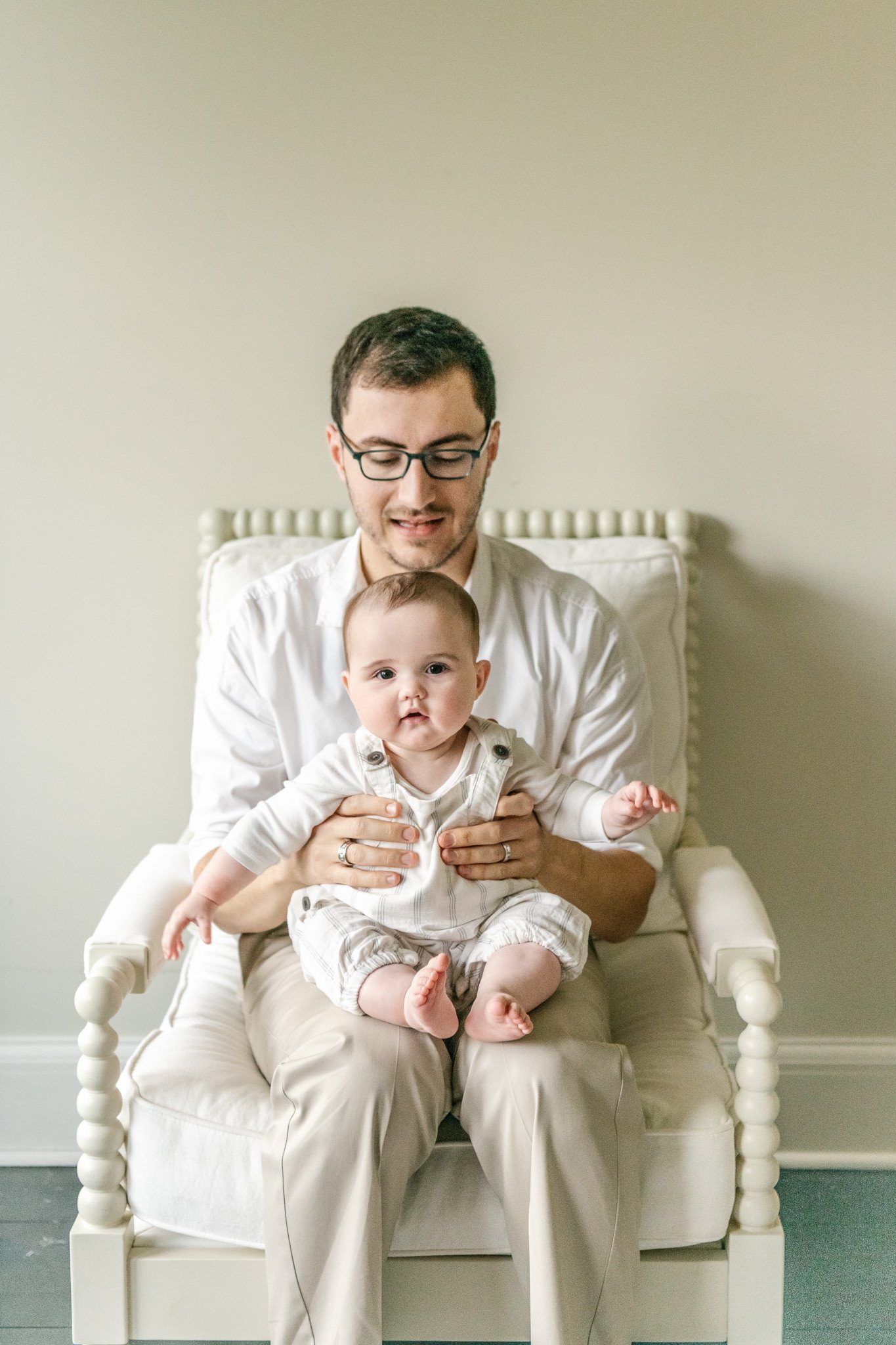  A baby girl sits on her daddy's lap during an at home family session with Nicole Hawkins Photography. white chair traditional timeless New Jersey #NicoleHawkinsPhotography #NicoleHawkinsfamilies #AtHomePortraits #EastCoastFamilyPhotographer #NewJers