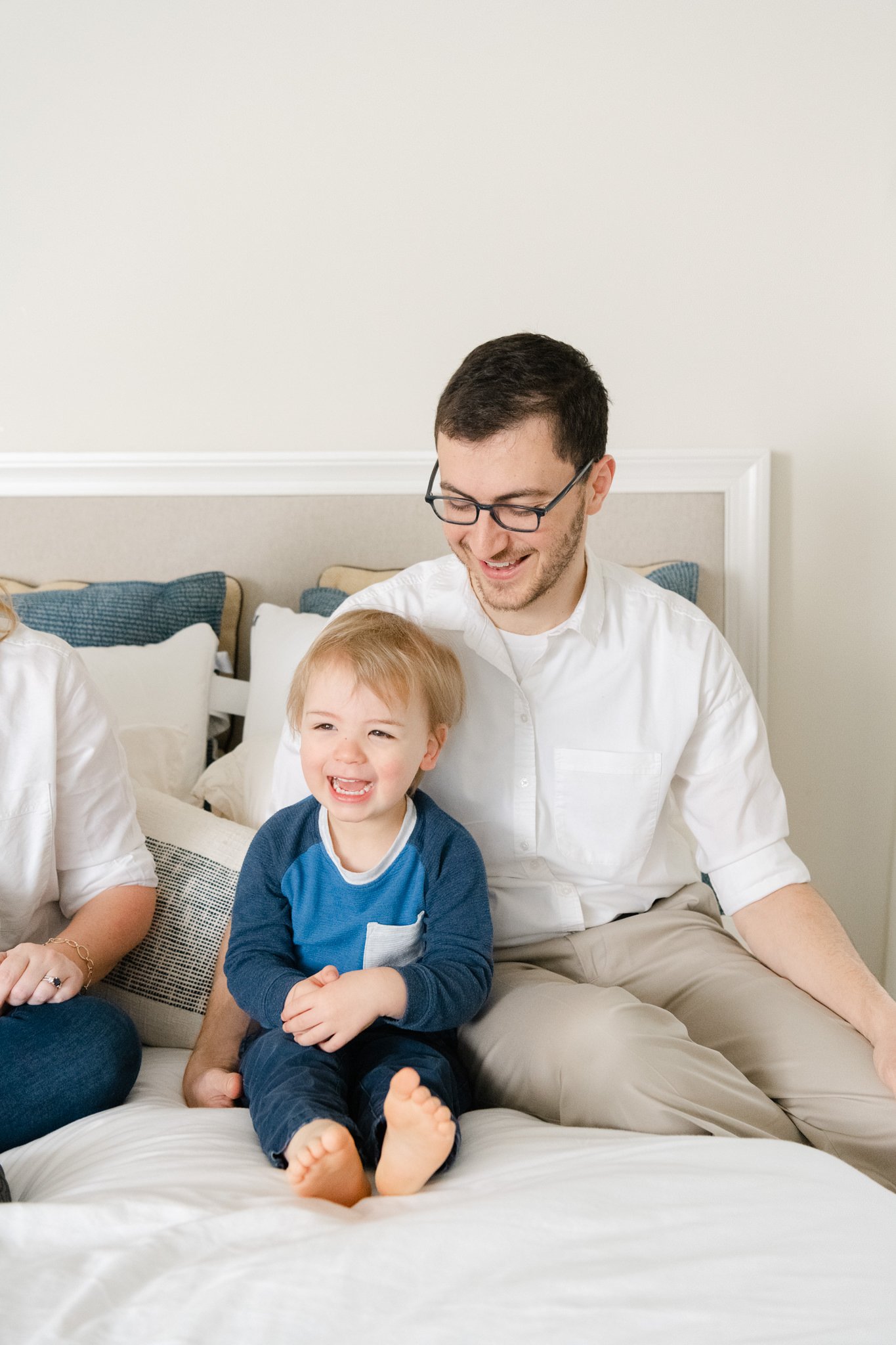  A dad with glasses smiles down at his little toddler boy while sitting on a bed by Nicole Hawkins Photography. toddler boy and dad sitting on bed #NicoleHawkinsPhotography #NicoleHawkinsfamilies #AtHomePortraits #EastCoastFamilyPhotographer #NewJers