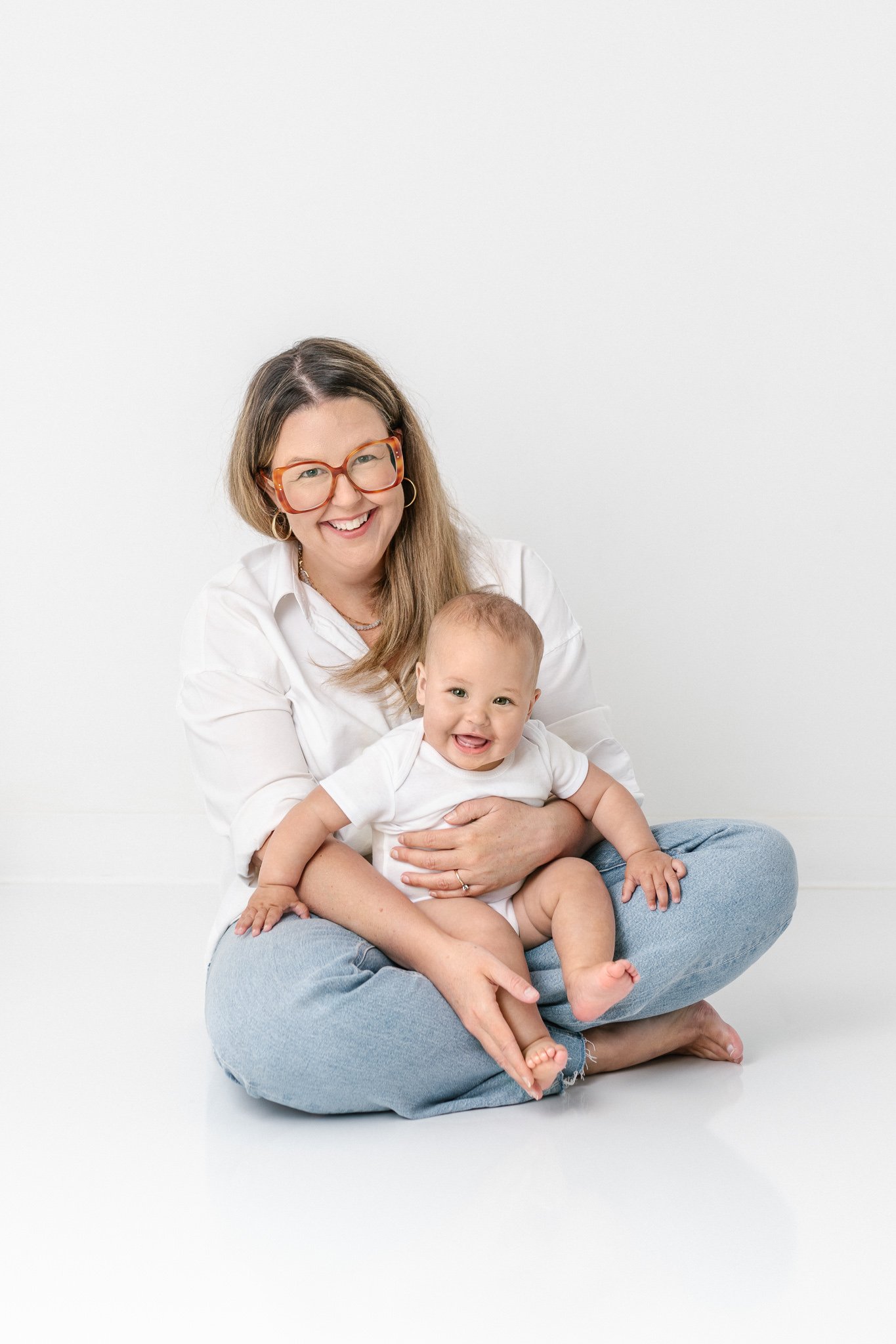  A mother wearing a white shirt and jeans holds her baby on her lap for a timeless, classic, modern portrait by Nicole Hawkins Photography. classic outfits timeless outfits #NicoleHawkinsPhotography #NicoleHawkinsNewborns #studionewborns #studioportr