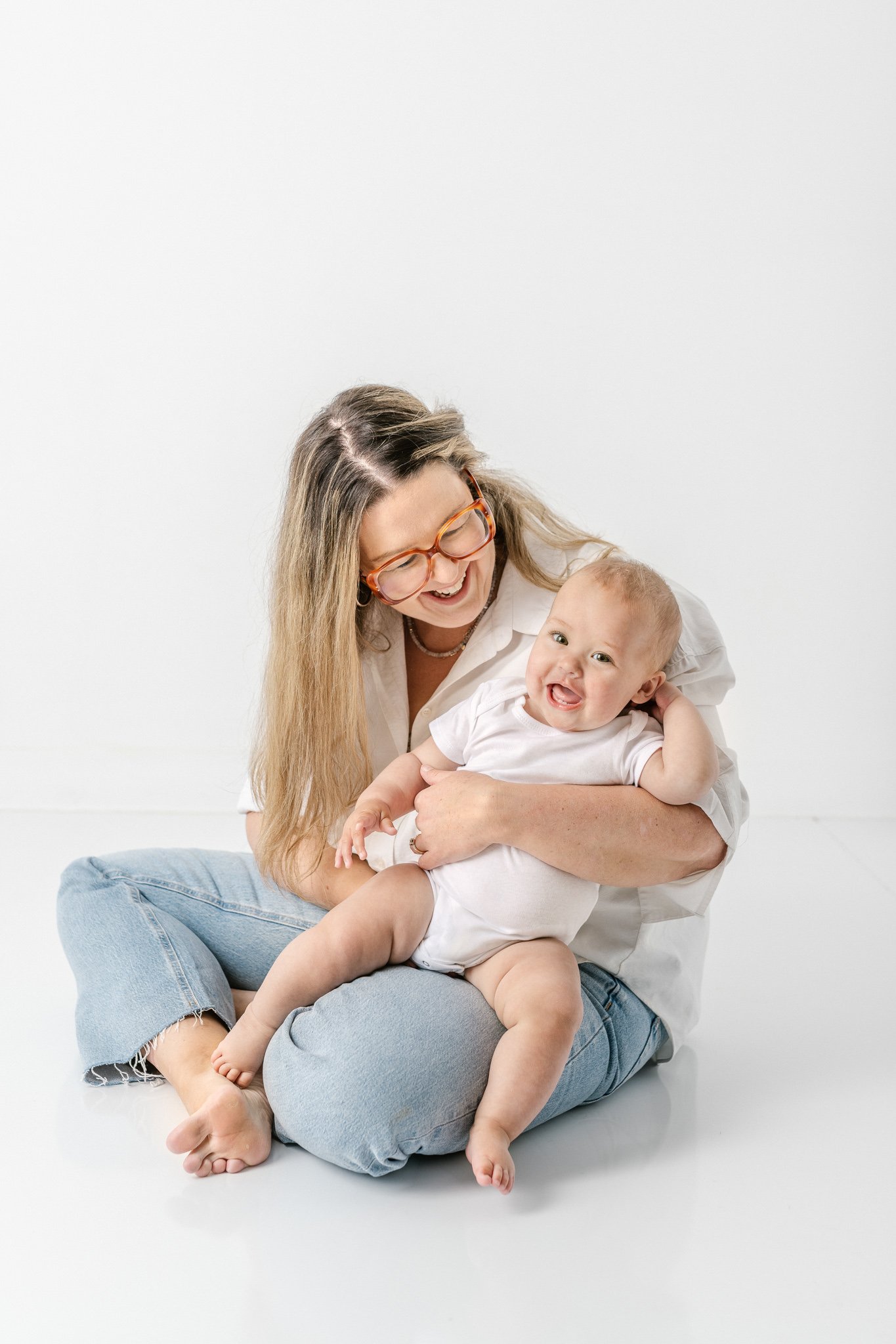  A momma holds her baby as her little girl laughs showing off her big toothy smile and chunky legs by Nicole Hawkins Photography. baby body toothy smile momma mom jeans #NicoleHawkinsPhotography #NicoleHawkinsNewborns #studionewborns #studioportraits