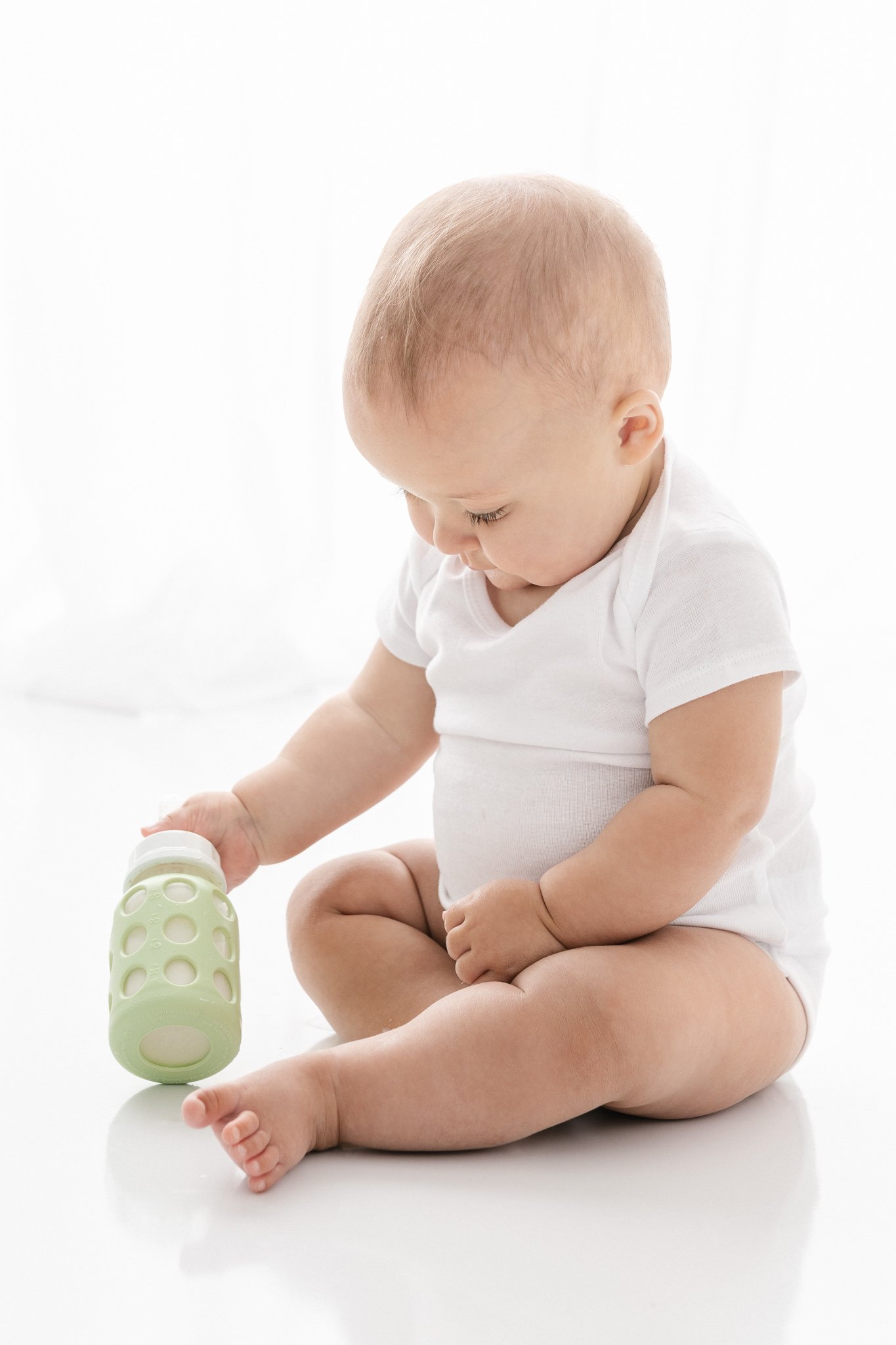  In a New Jersey studio, a baby plays with her bottle while wearing a white onesie by Nicole Hawkins Photography. baby playing with bottle green bottle #NicoleHawkinsPhotography #NicoleHawkinsNewborns #studionewborns #studioportraits #babystudioportr