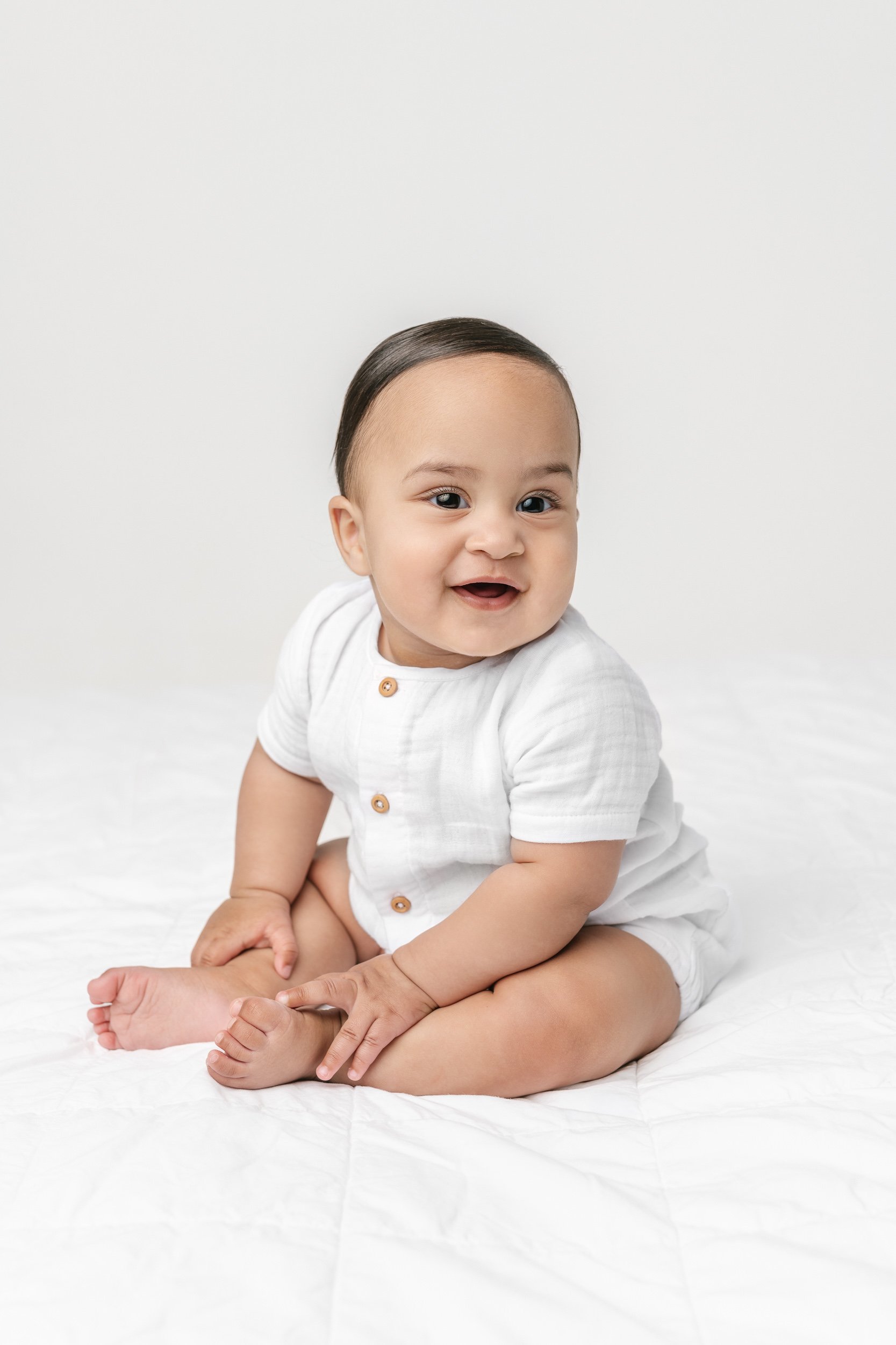  A one-year-old baby boy sits and smiles in a white romper captured by Nicole Hawkins Photography. white baby romper portraits NJ studio family #NicoleHawkinsPhotography #NicoleHawkinsNewborns #NewJerseyNewborns #NewJerseyStudioNewborns #StudioNewbor
