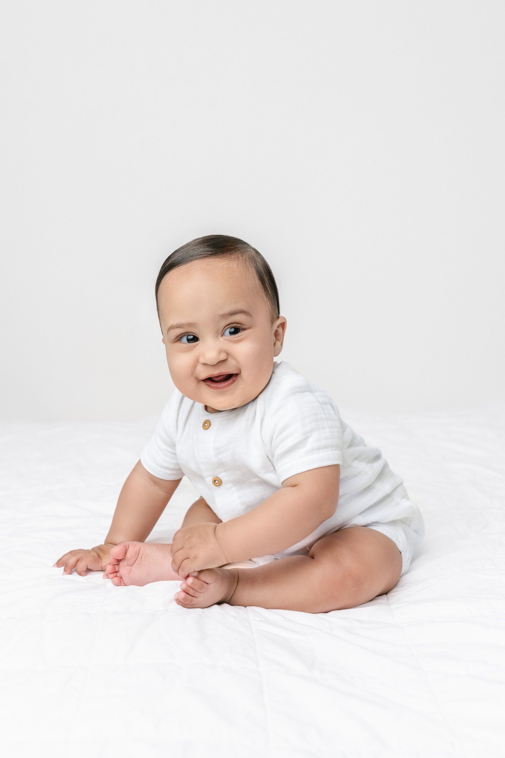  A beautiful chunky baby boy smiles while sitting on a bed in a studio on the east coast with Nicole Hawkins Photography. Stunning brown-eyed baby #NicoleHawkinsPhotography #NicolewHawkinsChildren #NewJerseyStudioBabies #StudioNewborn #StudioFamily #