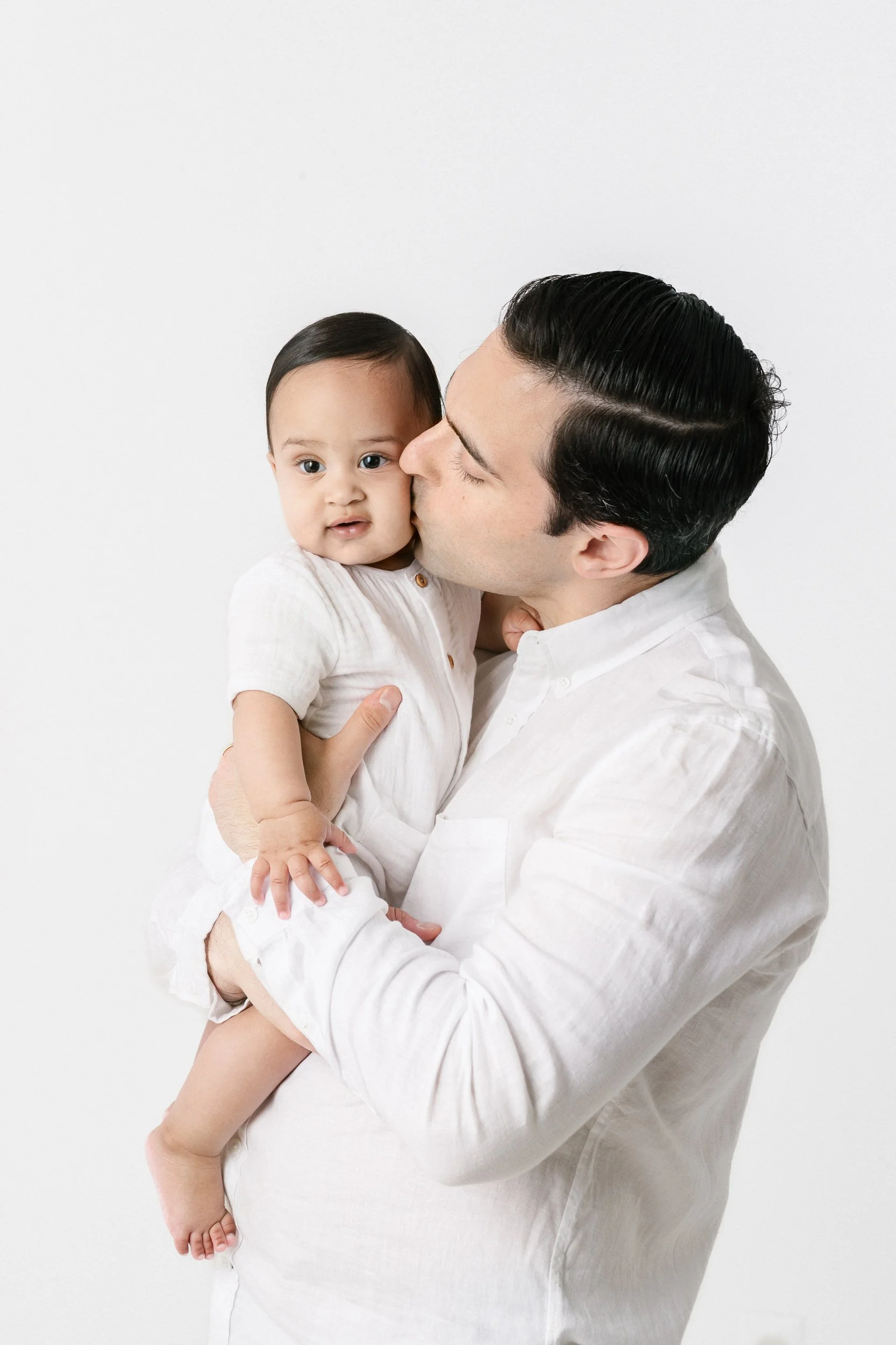  A father kisses his son on the cheek while holding him in his arms by Nicole Hawkins Photography. daddy and baby boy father kisses chunky baby #NicoleHawkinsPhotography #NicolewHawkinsNewborns #NewJerseyNewborns #NewJerseyStudioNewborns #StudioNewbo