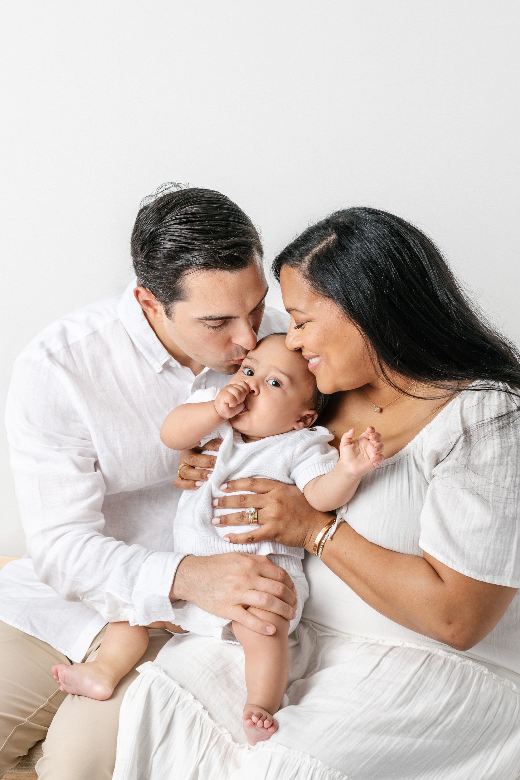  A chunky little boy sucks his thumb while mom and dad snuggle and kiss him by Nicole Hawkins photography. only child portraits snuggle mommy and daddy portraits #NicoleHawkinsPhotography #NicolewHawkinsNewborns #NewJerseyNewborns #NewJerseyStudioNew