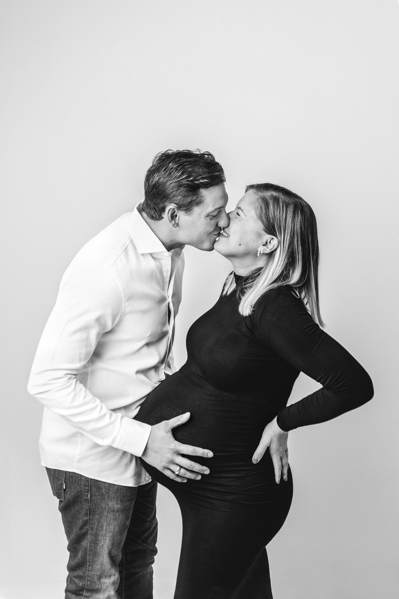  A husband holds his wife's baby bump while giving her a big kiss by Nicole Hawkins Photography. parents to be portrait studio maternity photography black maternity dress #NicoleHawkinsPhotography #NicoleHawkinsMaternity #MaternityStudioPortraits #Fa