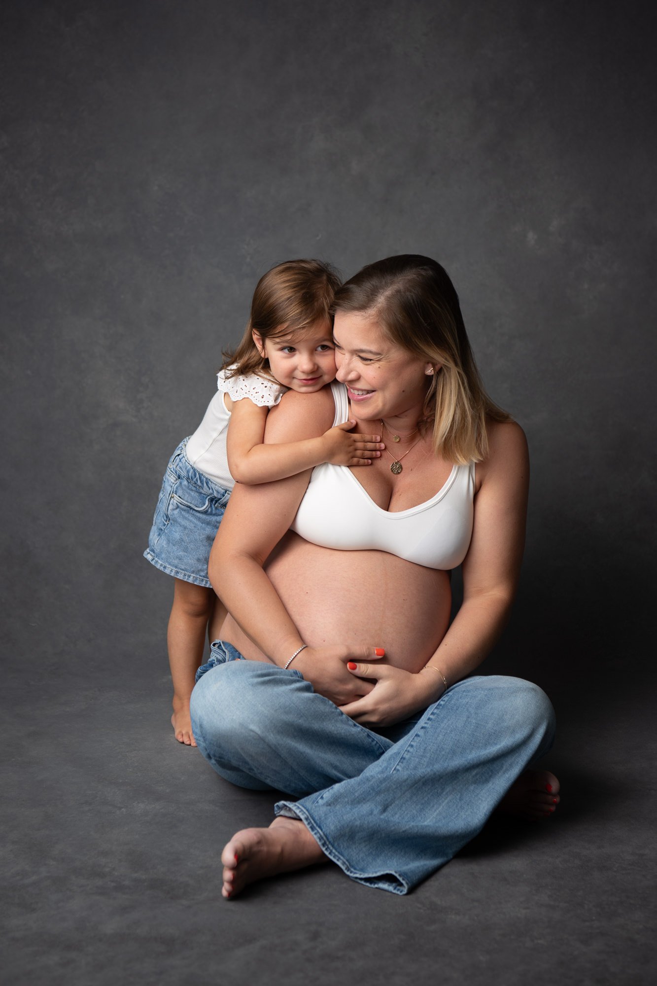  A mother sits with her legs crossed and pregnant belly in between while her little girl hugs her from behind by Nicole Hawkins Photography. mother daughter poses with pregnant belly #NicoleHawkinsPhotography #NicoleHawkinsMaternity #MaternityStudioP