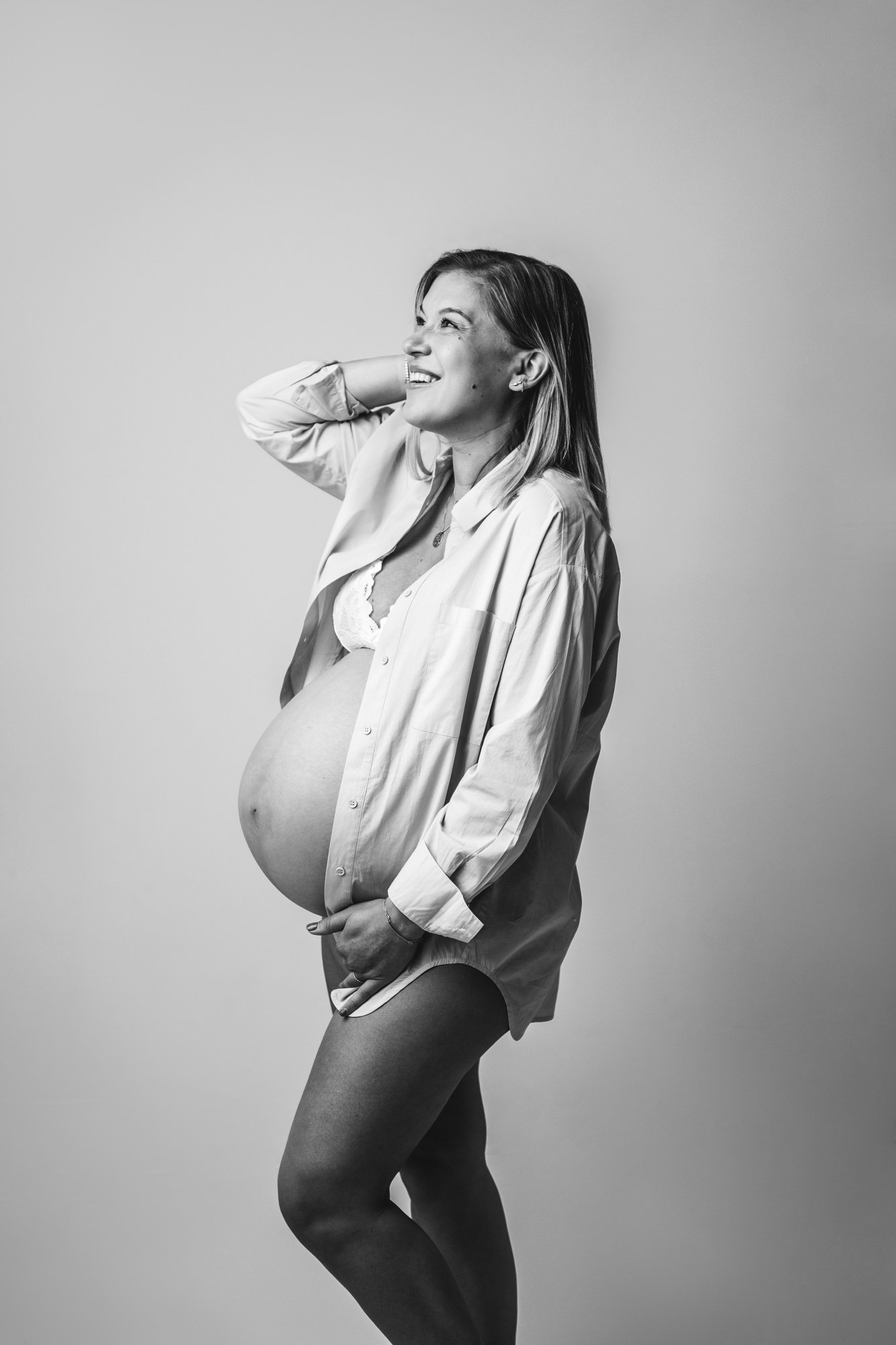  A black and white portrait of a pregnant woman with an exposed belly and button-up shirt by Nicole Hawkins Photography. exposed belly with button up shirt and bra #NicoleHawkinsPhotography #NicoleHawkinsMaternity #MaternityStudioPortraits #FamilyMat