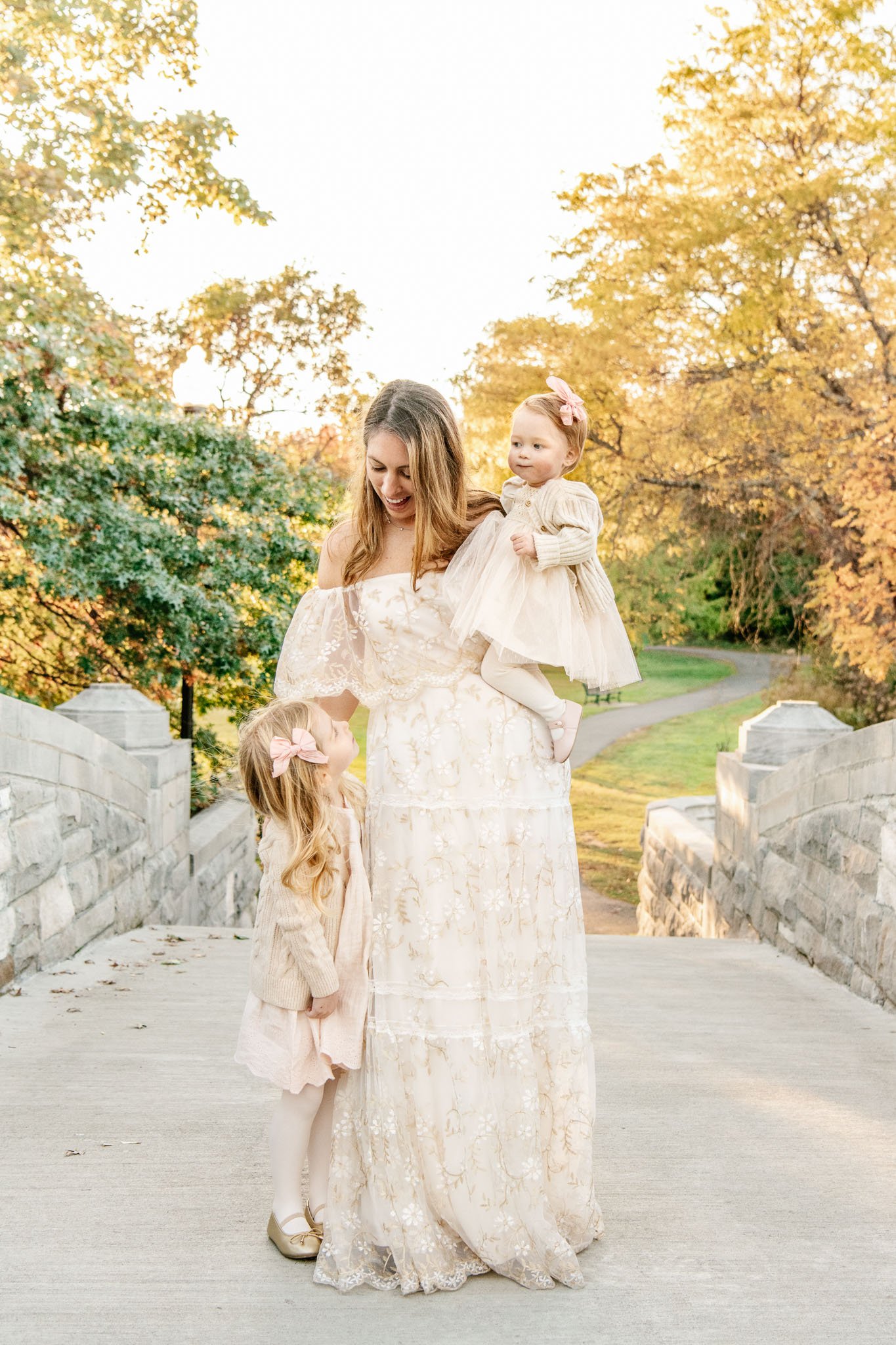  A mother in a white lacey dress smiles at her young daughters, captured by Nicole Hawkins Photography in NJ. Family with two little girls family wearing white for a family pic outside #NicoleHawkinsPhotography #NicoleHawkinsFamilies #FallFamilyPhoto