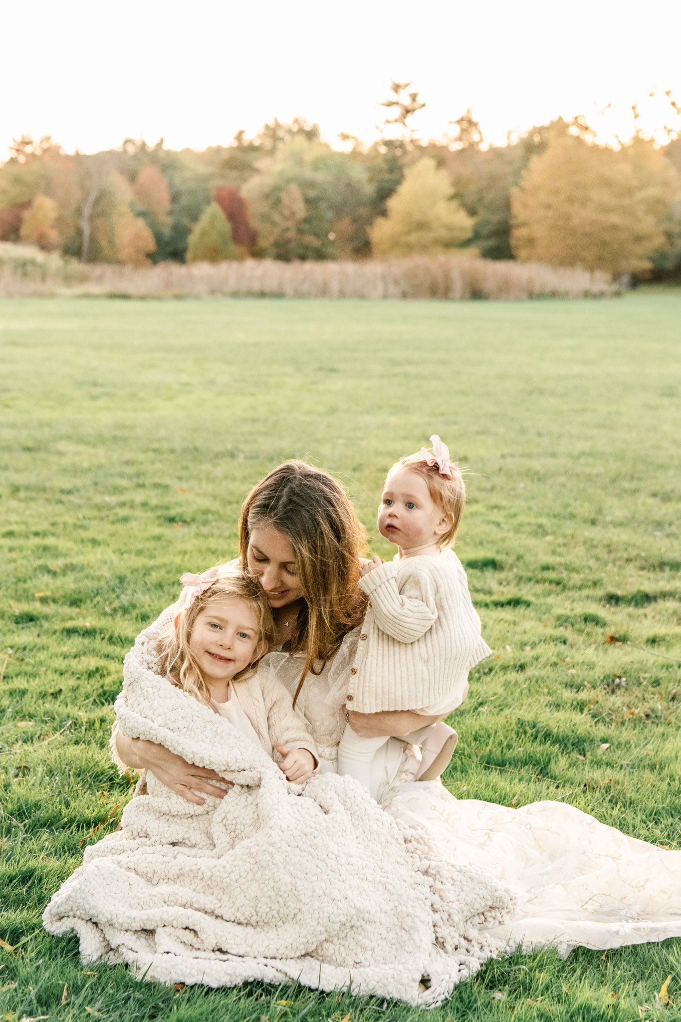  A mother sits on a grass field while holding two little girls with fall leaves behind them by Nicole Hawkins Photography. fall leaf family portrait  #NicoleHawkinsPhotography #NicoleHawkinsFamilies #FallFamilyPhotos #FallPortraits #NJFamilyPhotograp