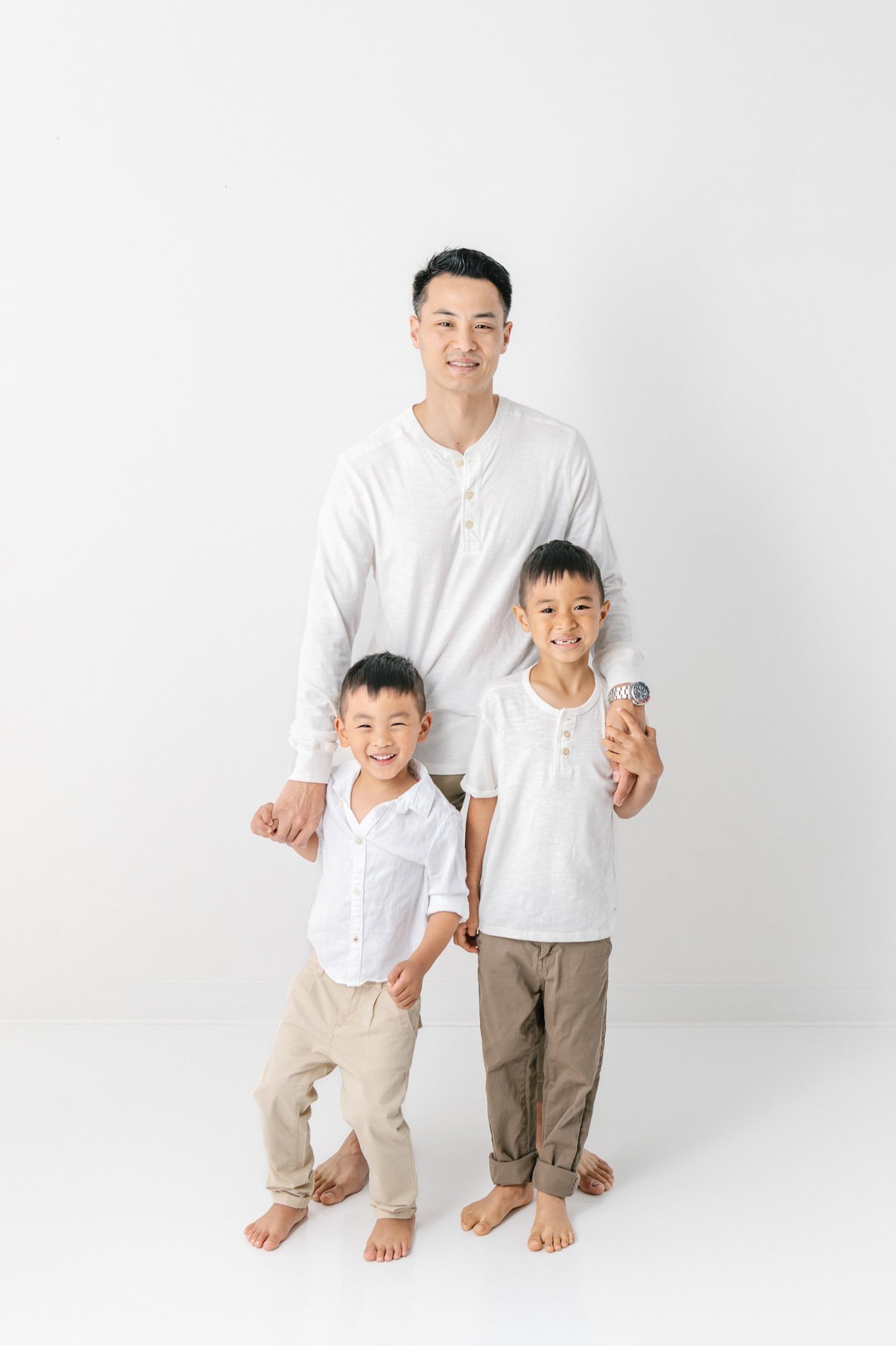    A dad and his two sons pose in a family photoshoot done by Nicole Hawkins Photography in New Jersey. Dad places an arm around both sons while everyone gazes at the camera. #studionewborns #studioportraits #babystudioportraits #NewJerseyPhotographe