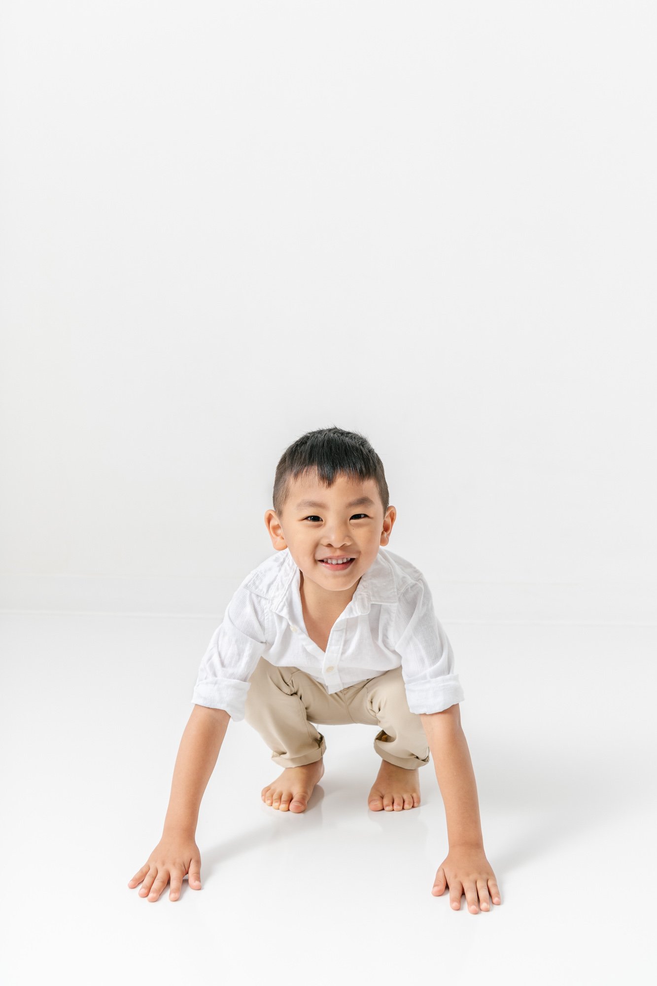    Young boy crouches down and touches the ground with a huge grin during studio photo session with Nicole Hawkins Photography. Family portraits in New Jersey #studionewborns #studioportraits #babystudioportraits #NewJerseyPhotographers #NJfamilyphot