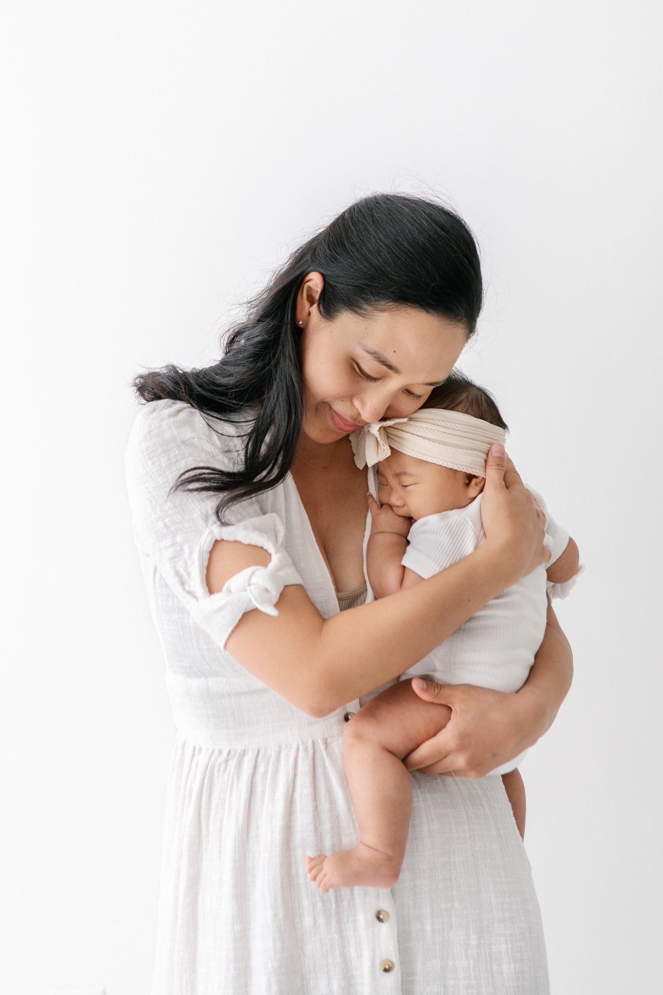    Young mother cradles newborn baby girl in white bow. Happy newborn and happy mother sharing a special moment in New Jersey shot by Nicole Hawkins Photography #studionewborns #studioportraits #babystudioportraits #NewJerseyPhotographers #NJfamilyph