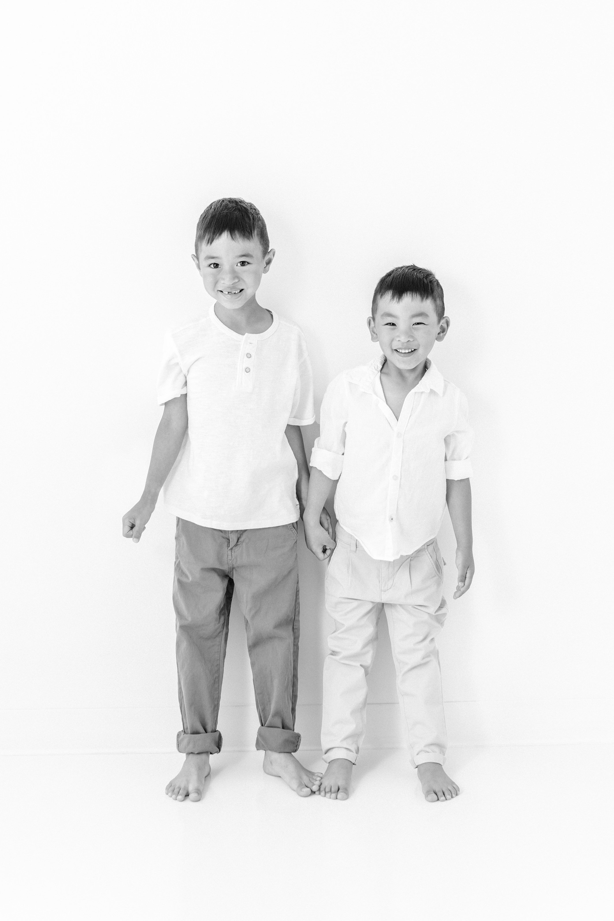   Two brothers shown in black and white studio portrait at a newborn family photo session in New Jersey. Pictures captured by Nicole Hawkins Photography. Brothers are both smiling wide at the camera while holding hands #studionewborns #studioportrai