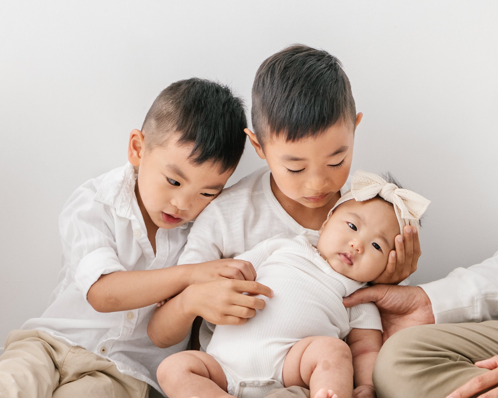    Siblings sit on the ground close together to admire newborn baby sister. Brothers hold hands and love on baby sister. Newborn baby looks at camera while family holds her. #studionewborns #studioportraits #babystudioportraits #NewJerseyPhotographer