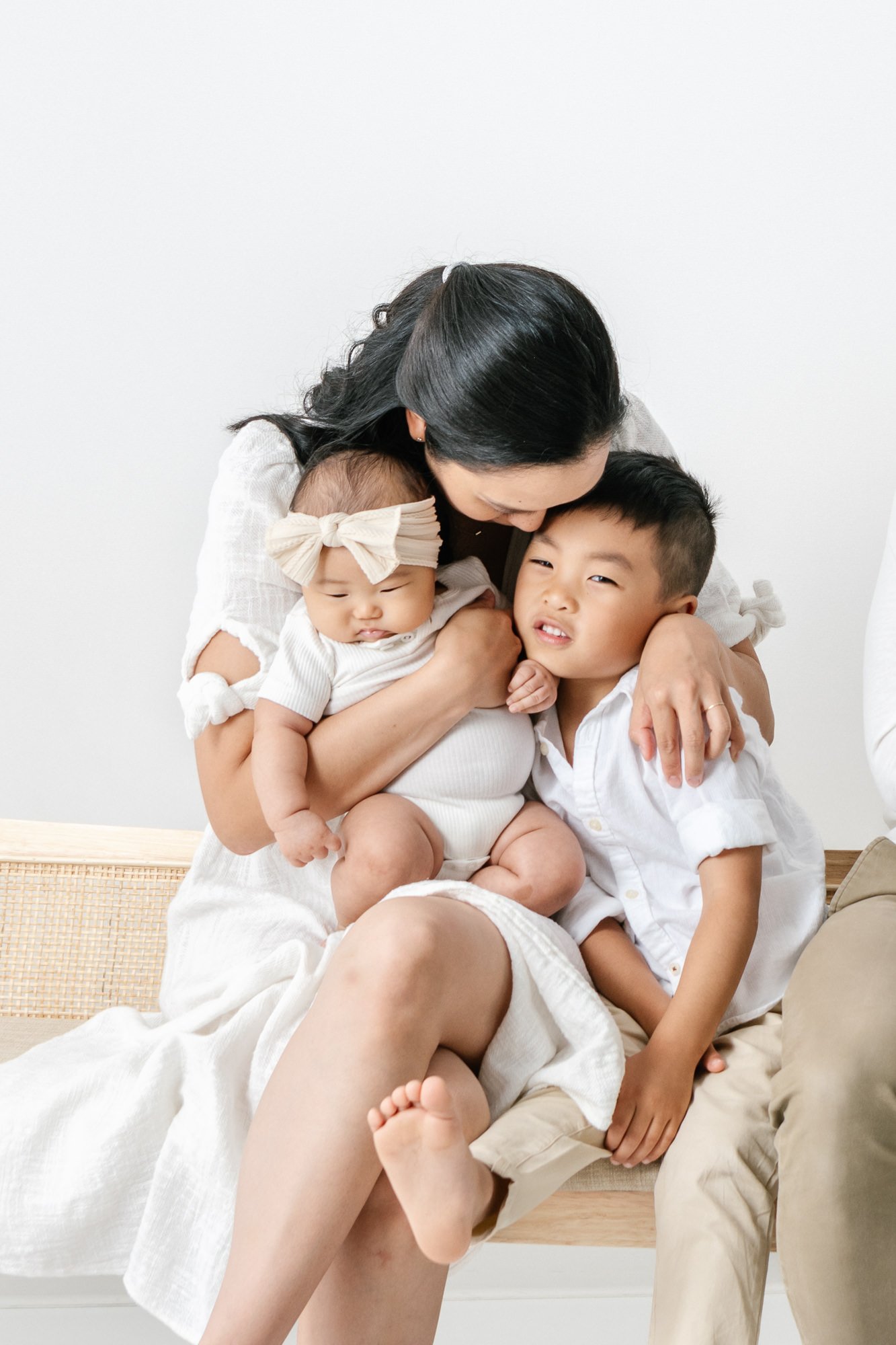    Sweet mother embraces two of her children. Holds baby on her lap and embraces son with one arm. All are dressed in white during Nicole Hawkins newborn photoshoot in New Jersey #studionewborns #studioportraits #babystudioportraits #NewJerseyPhotogr