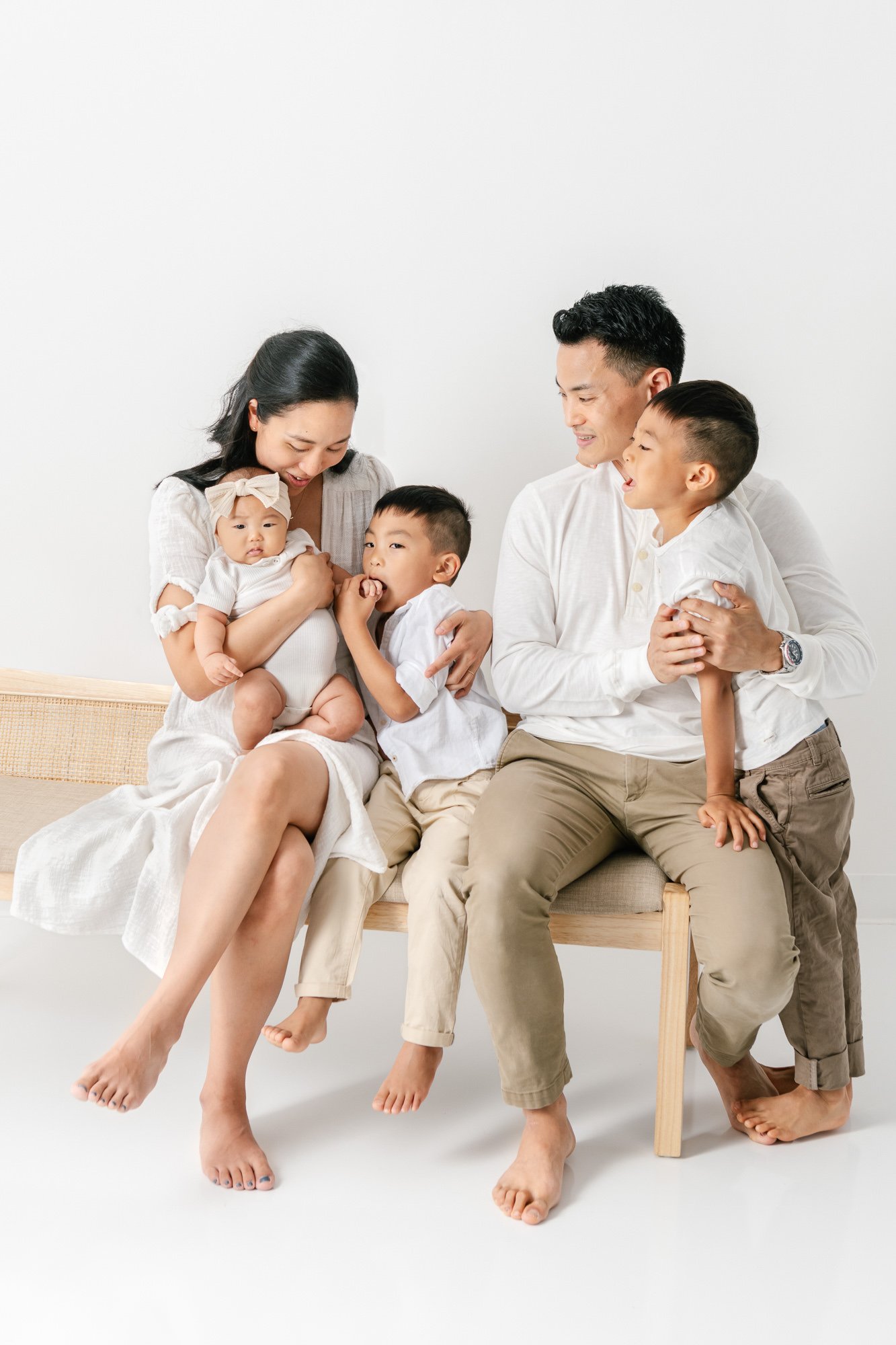    Family sits on a bench tucked in close together. Baby sister looks directly at camera. Brothers look to parents. Dad places arm around one son in newborn family photoshoot captured by Nicole Hawkins Photography #studionewborns #studioportraits #ba