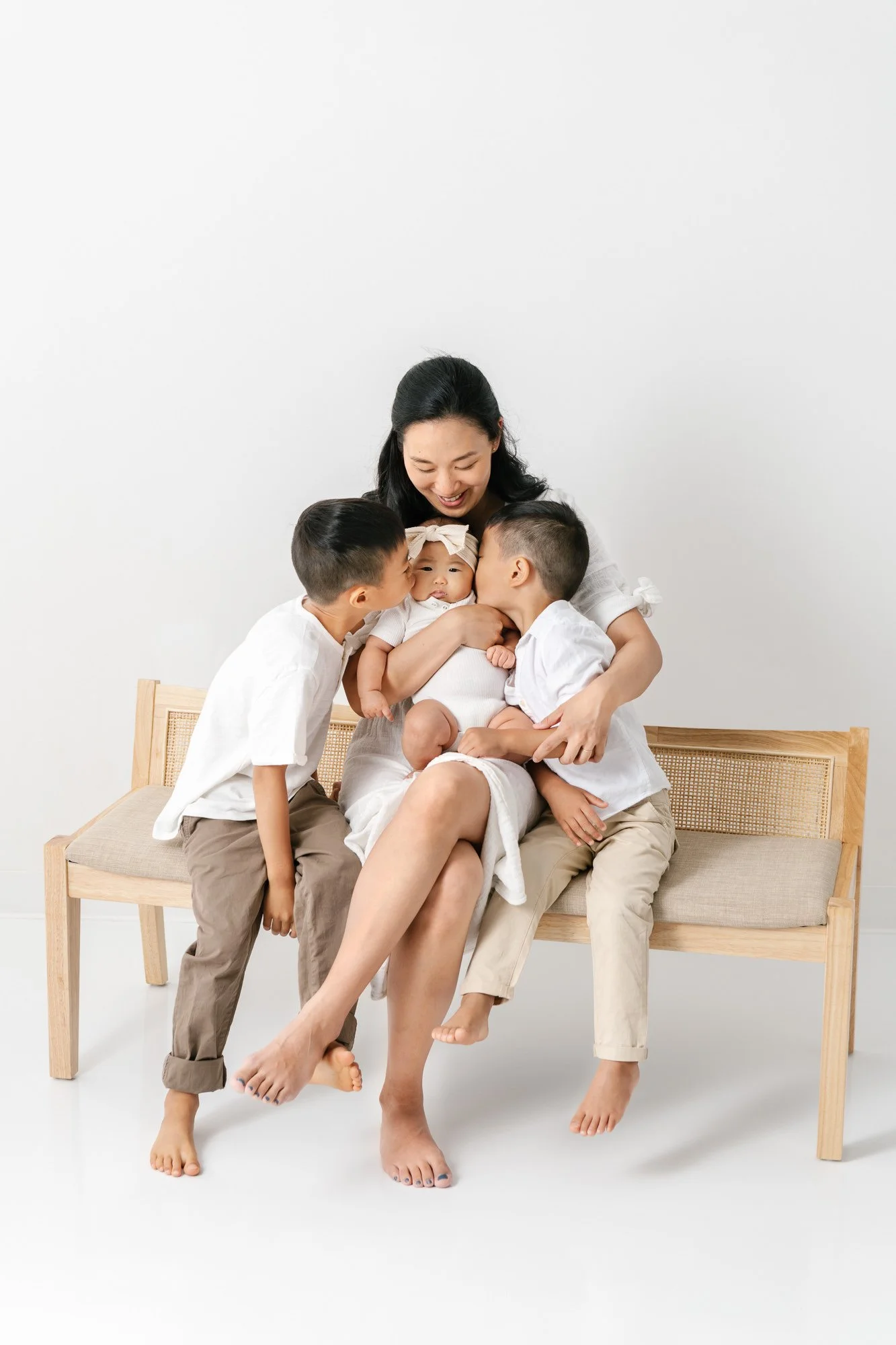    Mother sits on a bench surrounded by her children. Brothers plant a kiss on baby sister’s cheeks. Mother looks down happily in New Jersey at her children by Nicole Hawkins Photography #studionewborns #studioportraits #babystudioportraits #NewJerse