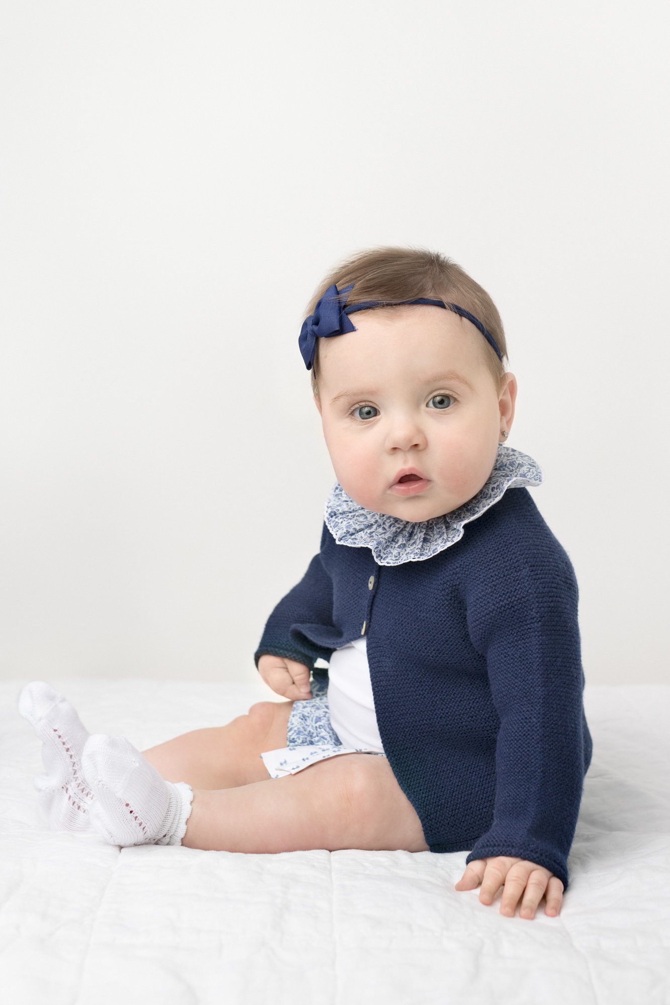  Young baby sitting up with a thoughtful look in an all white studio captured by Nicole Hawkins Photography in New Jersey area. Baby is wearing socks and a darling blue sweater with a white ruffle collar #babystudioportraits #studionewborns #NewJerse